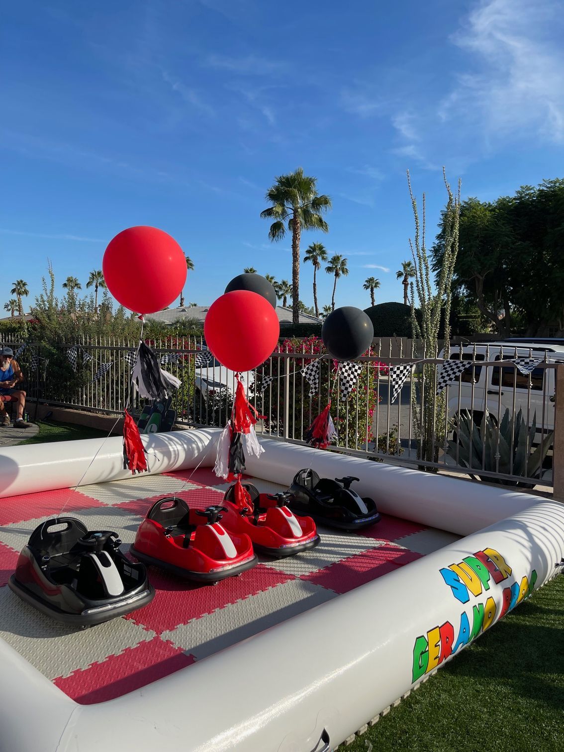 Mini race cars on a track with red and black balloons under a sunny blue sky