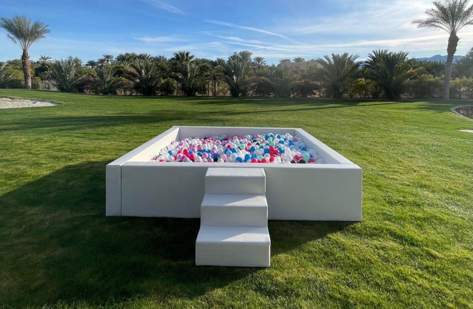 White ball pit outdoors on a grassy lawn with palm trees in the background.