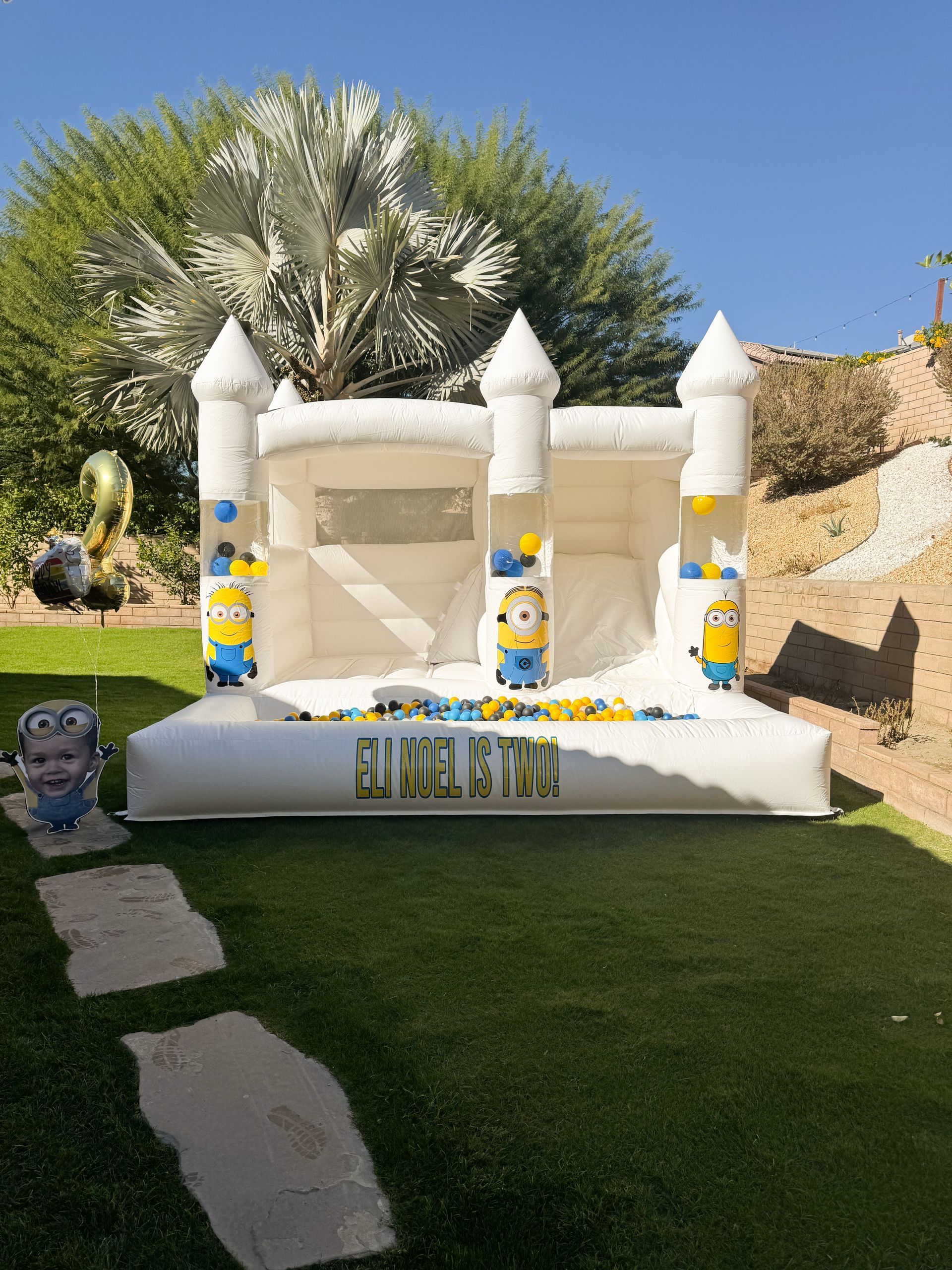 Minion-themed bounce house on a lawn with white castle turrets and a poolside backdrop
