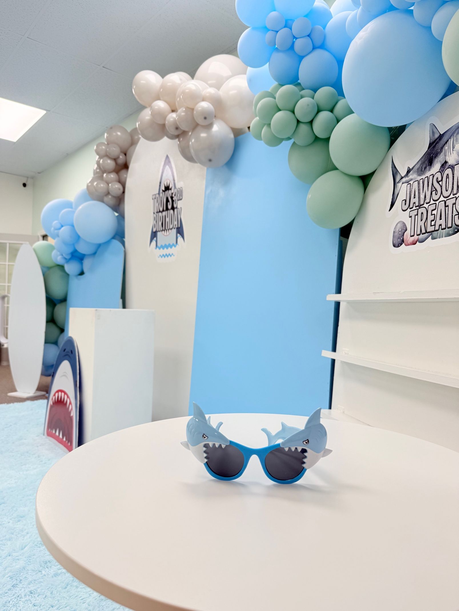 Pastel blue party backdrop with balloon garlands and a round white table with a small cake in front