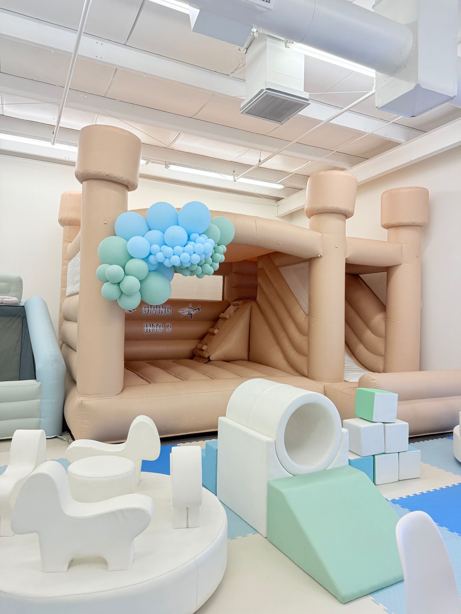 Indoor play area with beige inflatable structures, blue balloon cluster, and soft climbing ramps.
