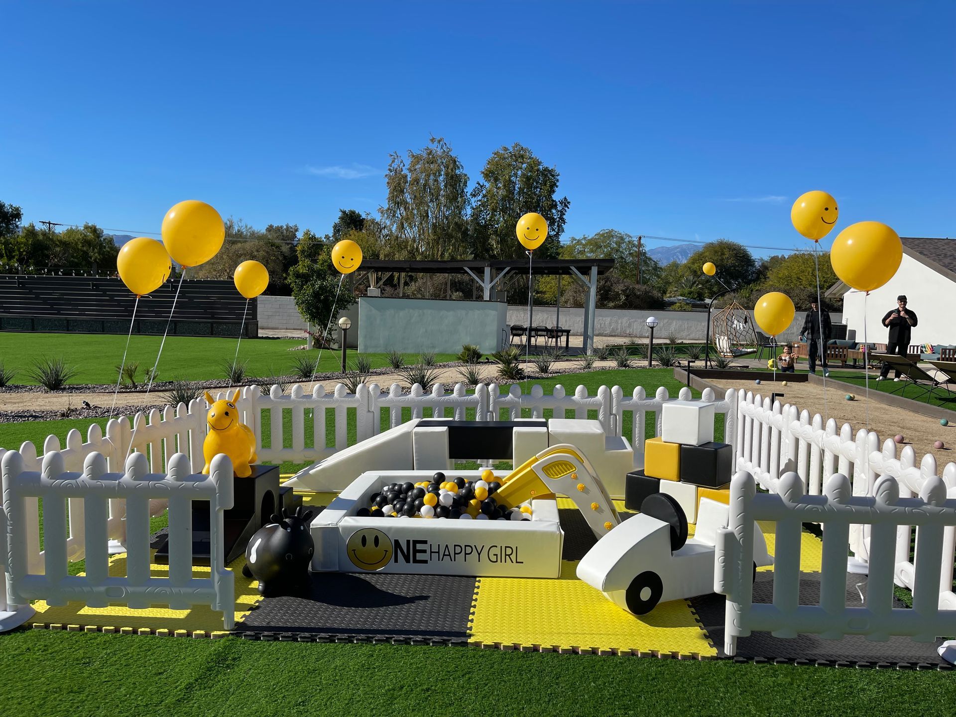 Yellow balloons decorate a fenced outdoor display with a small model car on grass under a blue sky