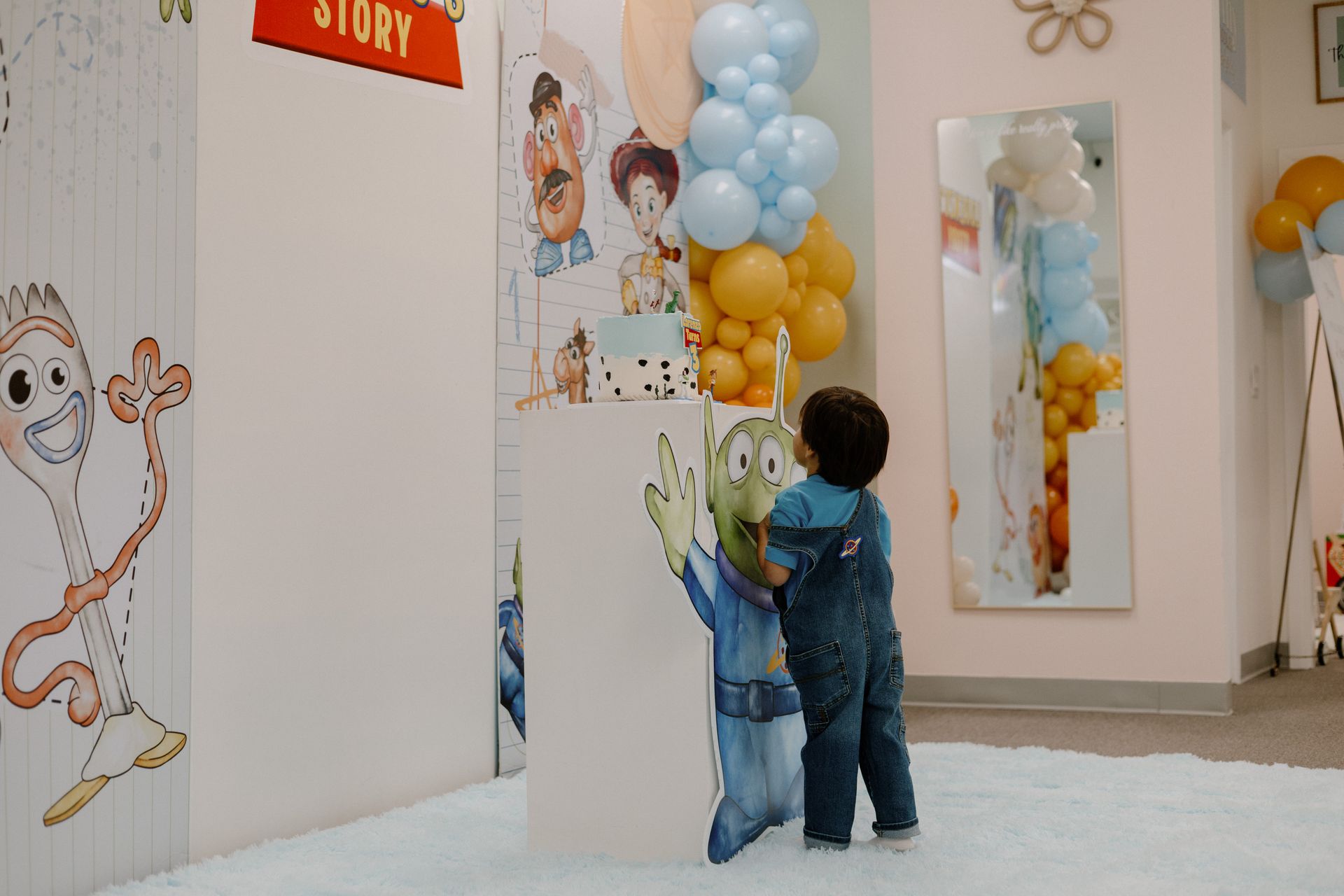 Child in denim overalls touches a cartoon cutout beside pastel balloons in a bright playroom