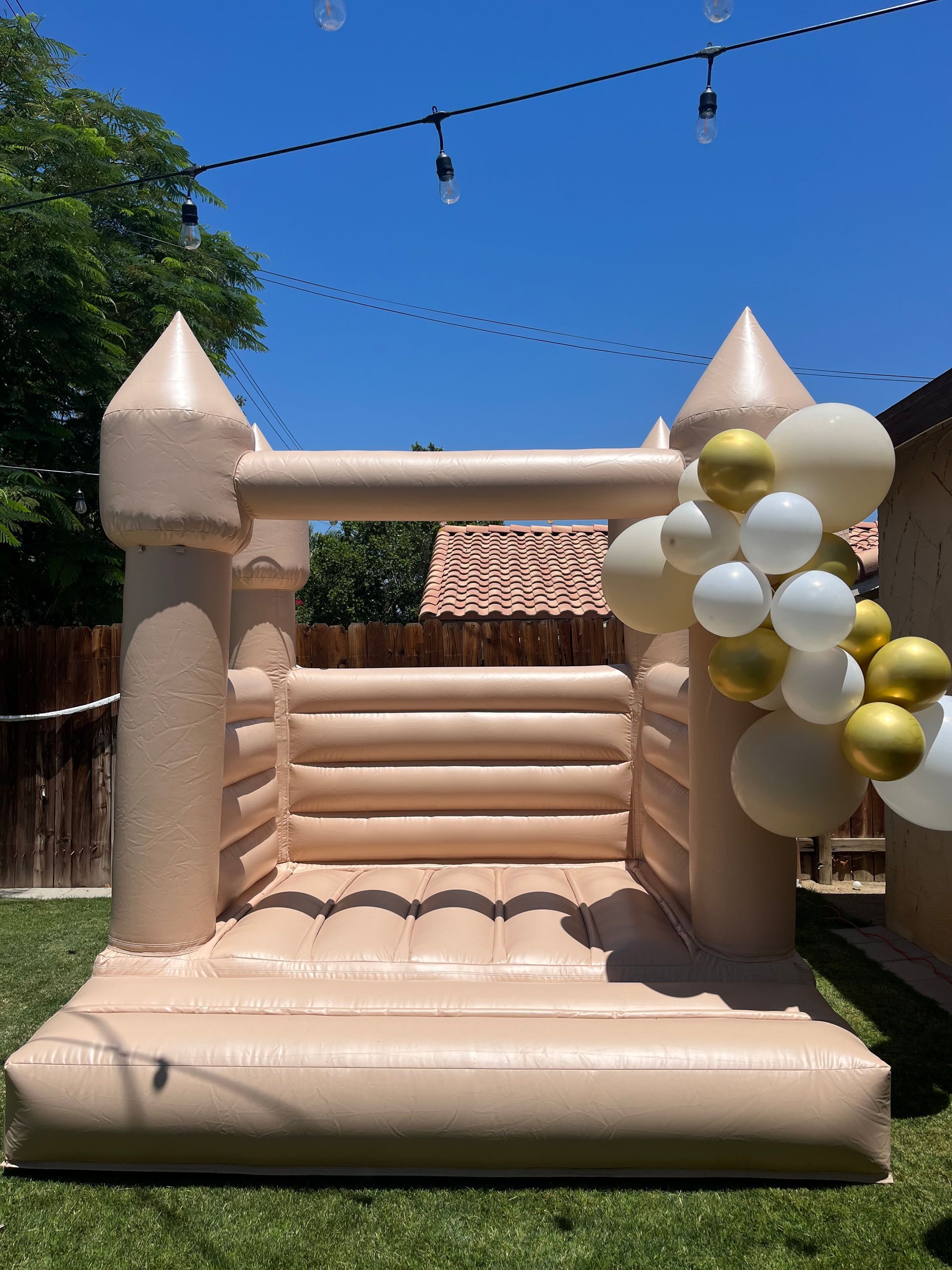 Beige inflatable bounce house with castle towers and gold balloons outdoors under a blue sky