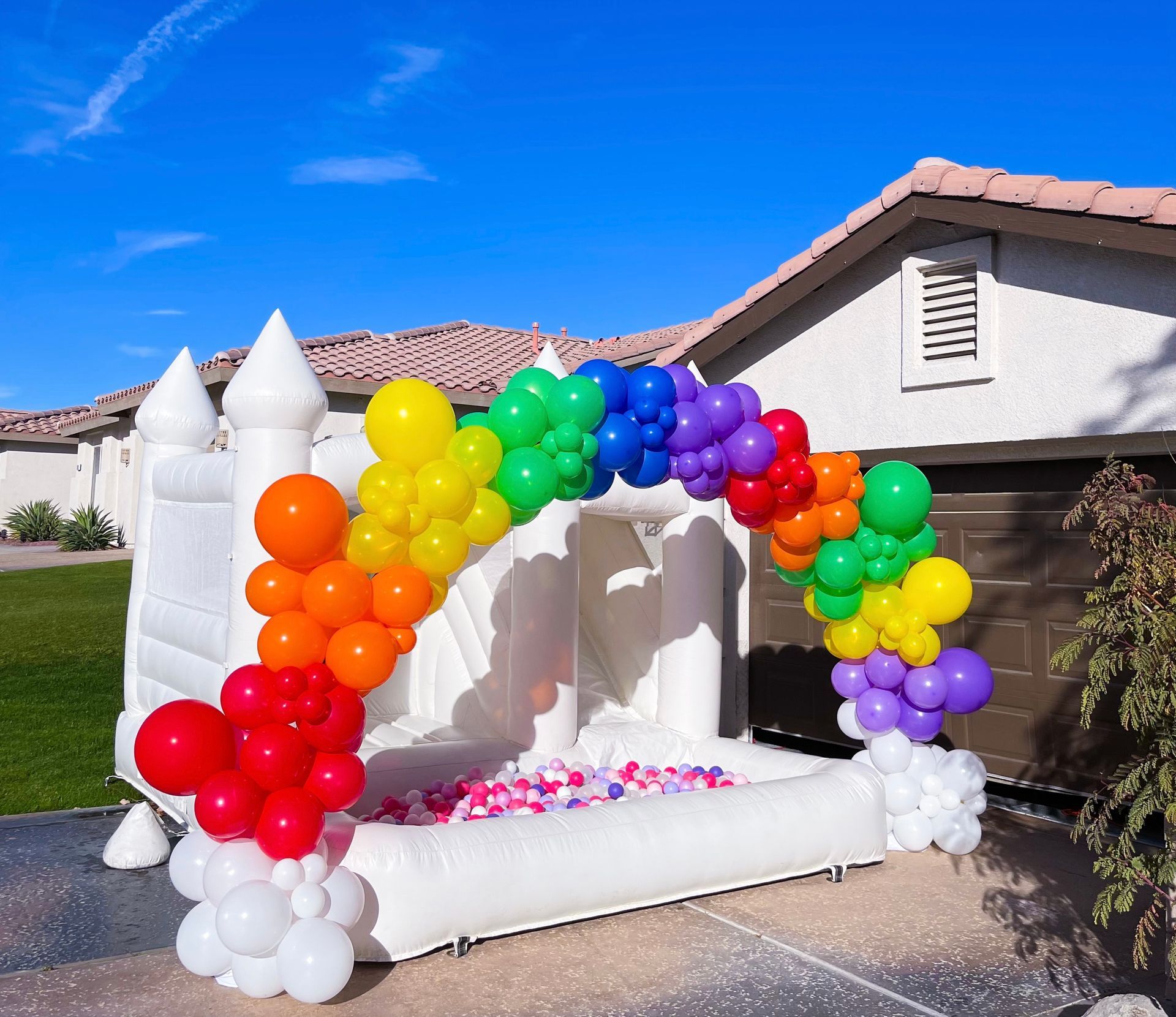 Colorful rainbow balloon arch around a white bounce house in a suburban driveway