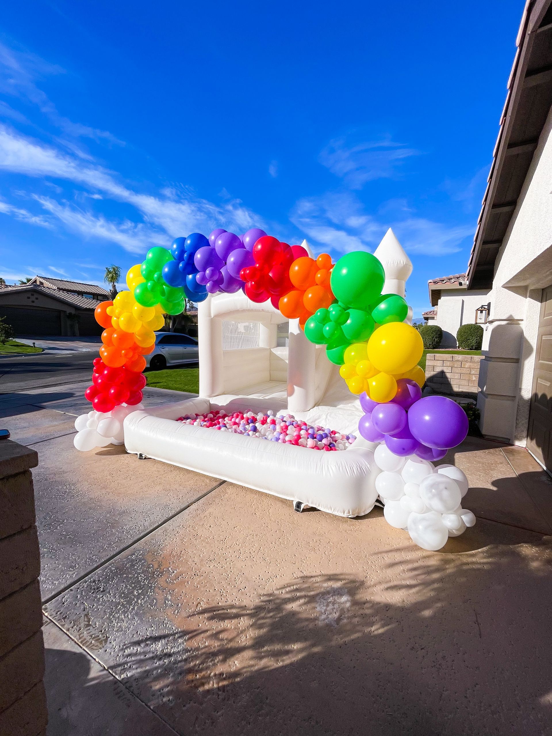 Rainbow balloon arch over a white outdoor bench with pink flowers in a sunny yard