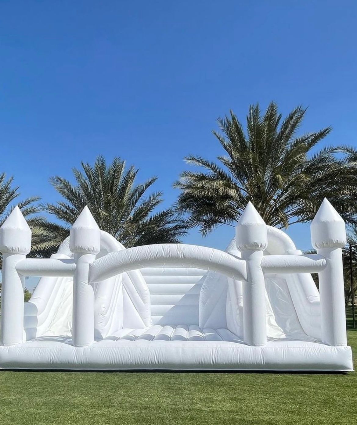 White inflatable bounce castle with turrets on grass under a bright blue sky, palm trees behind