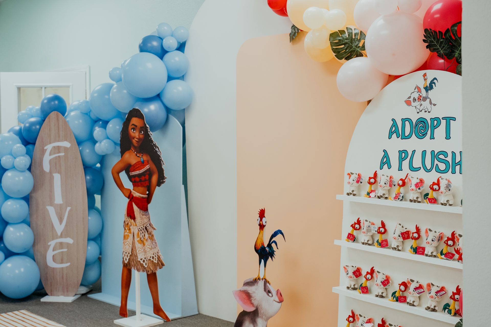 Outdoor birthday display with blue and white balloons, surfboard, doll cutout, and “Adopt a Plush” shelf