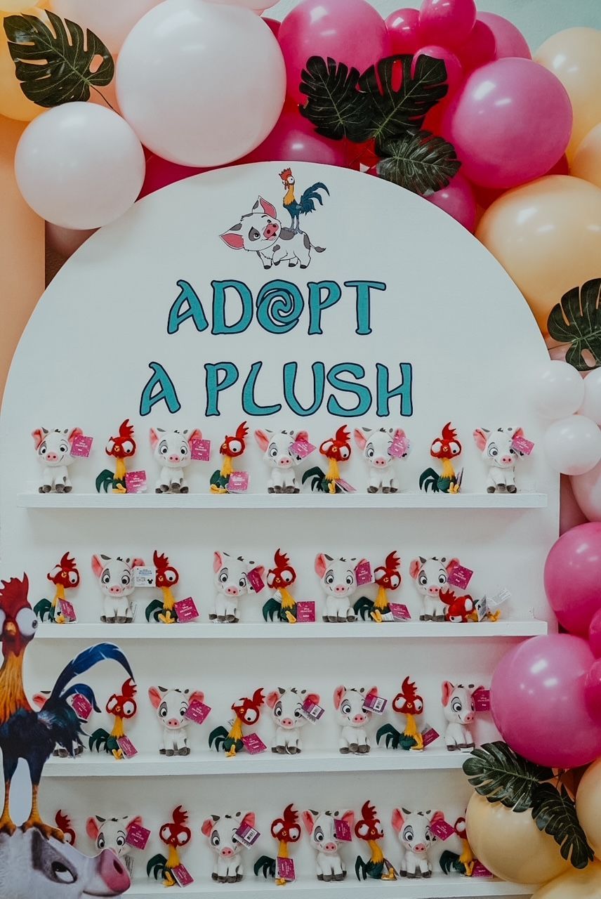 “Adopt a Plush” display with white shelves of small plush toys, pink and white balloons, and leaves at the top.