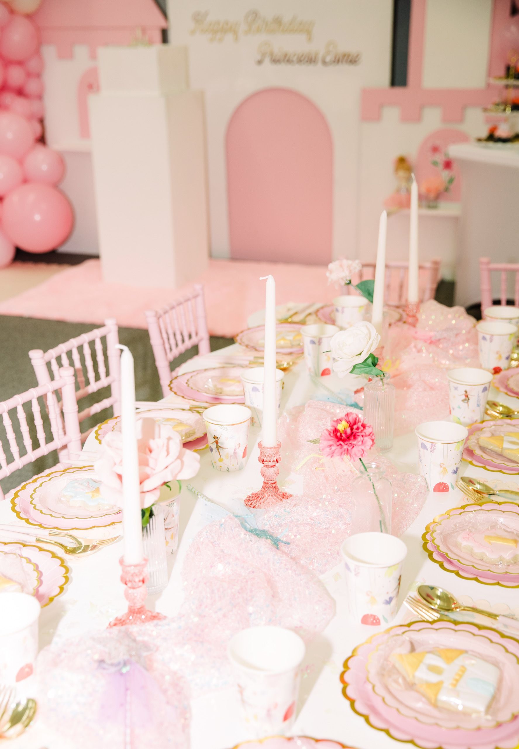 Pink dessert party table with floral decor, white chairs, and pastel arch backdrop