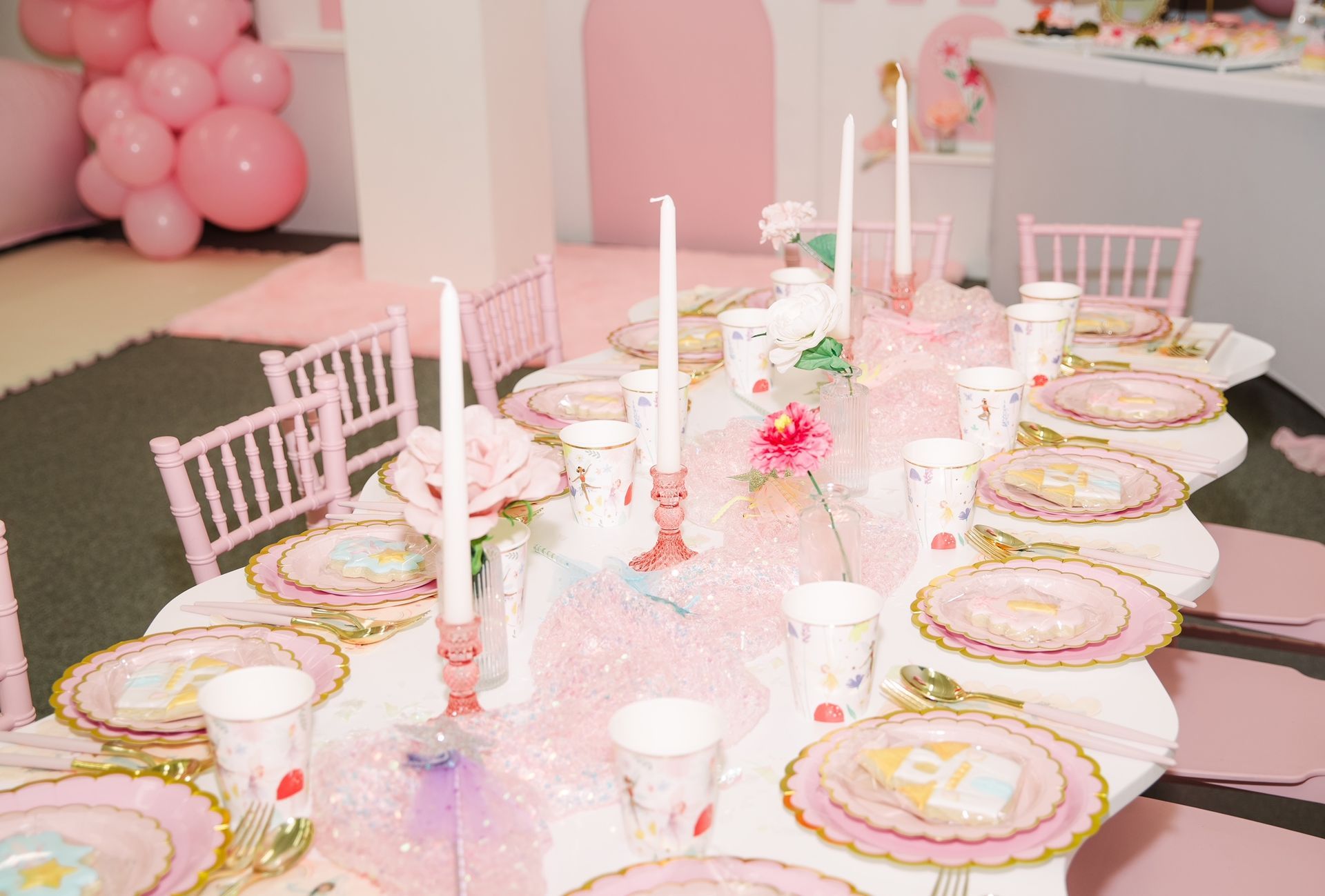 Pink dessert table set for a tea party with candles, plates, cups, and balloons in the background