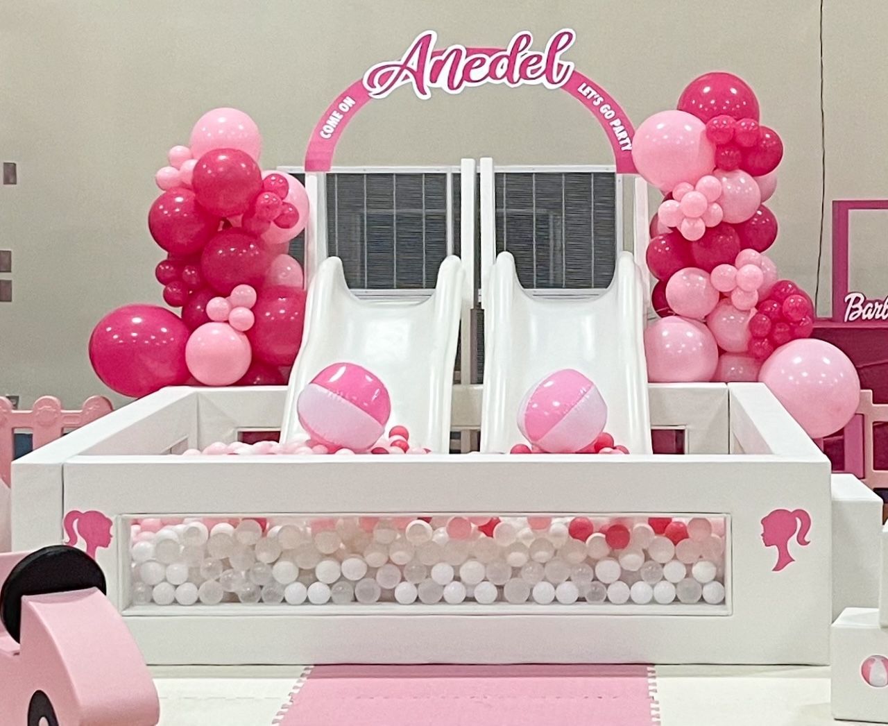 Pink balloon-decorated indoor play area with twin slides and a name arch over a white ball pit