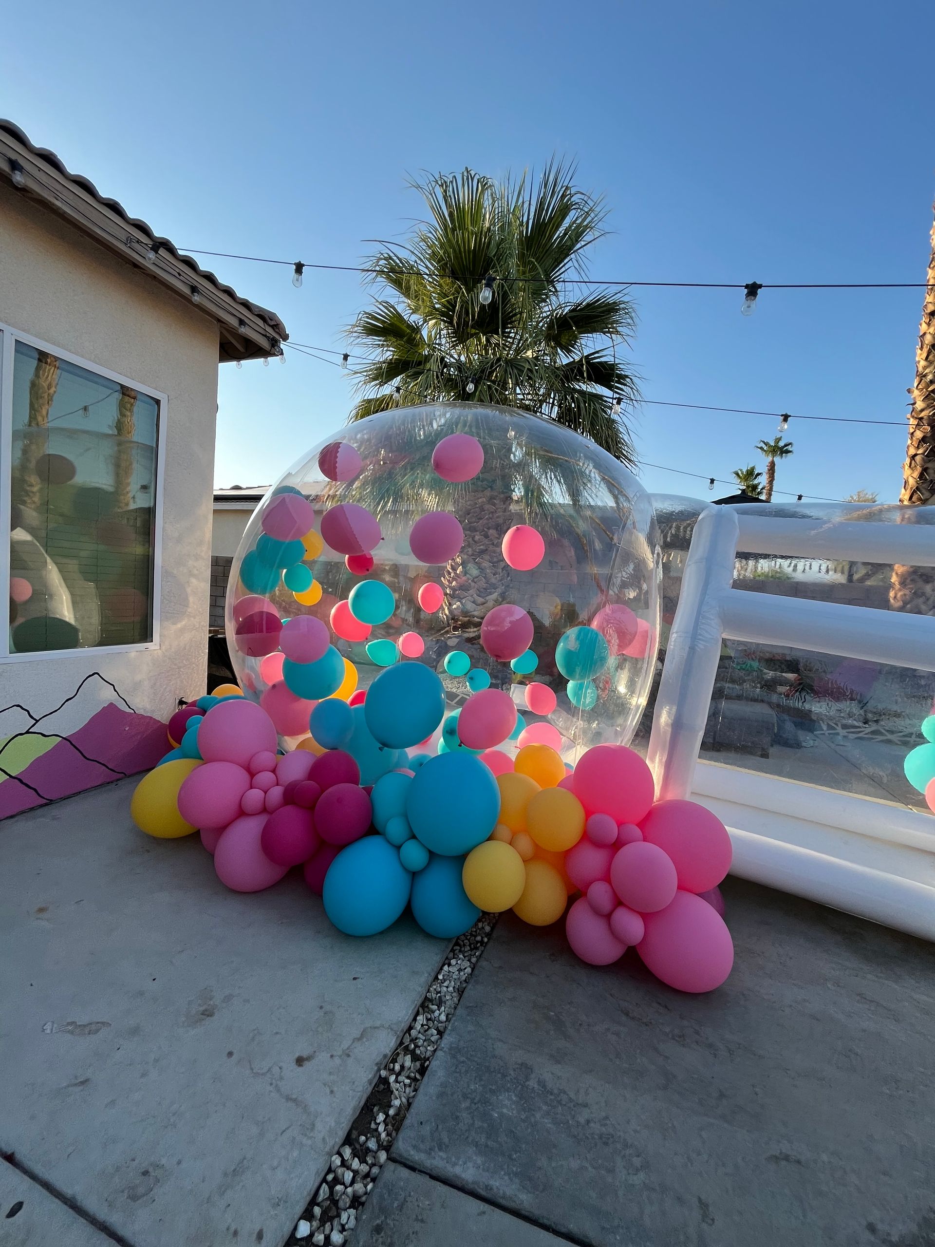 Clear inflatable dome filled with colorful balloons on a patio beside a house, under string lights.