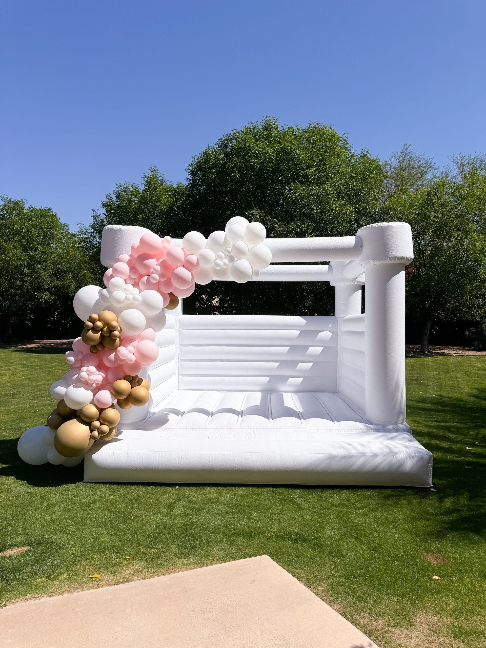 White inflatable bounce house decorated with pink, white, and gold balloon clusters on a sunny lawn
