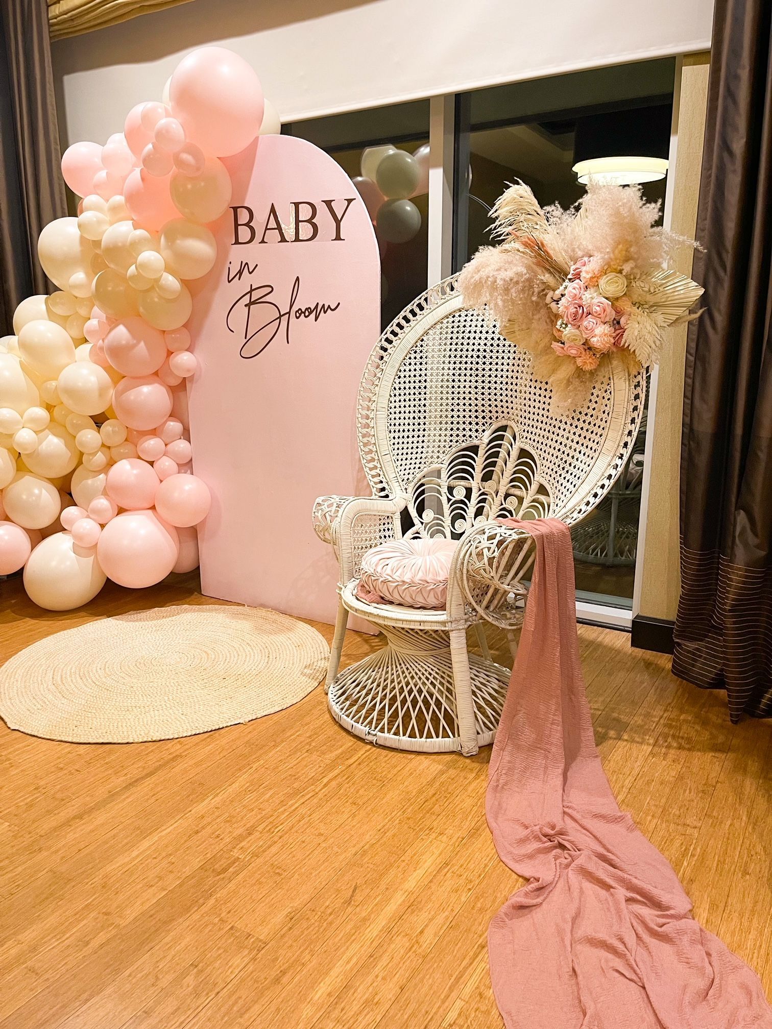 Baby shower decor with pink balloons, floral sign, white wicker chair, and pink draped aisle on a wooden floor.
