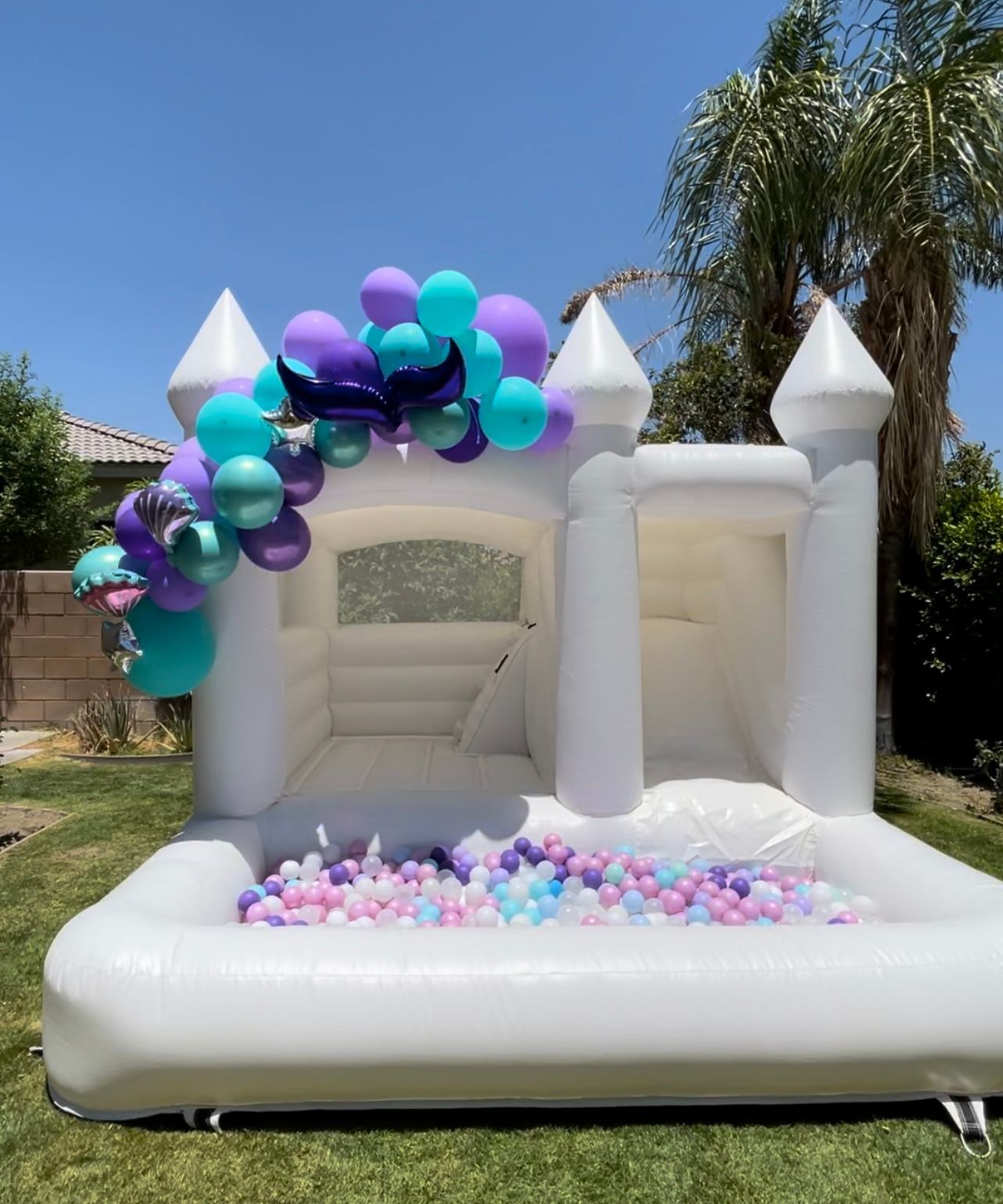 White inflatable bounce house with purple, teal, and pink balloons in a sunny backyard