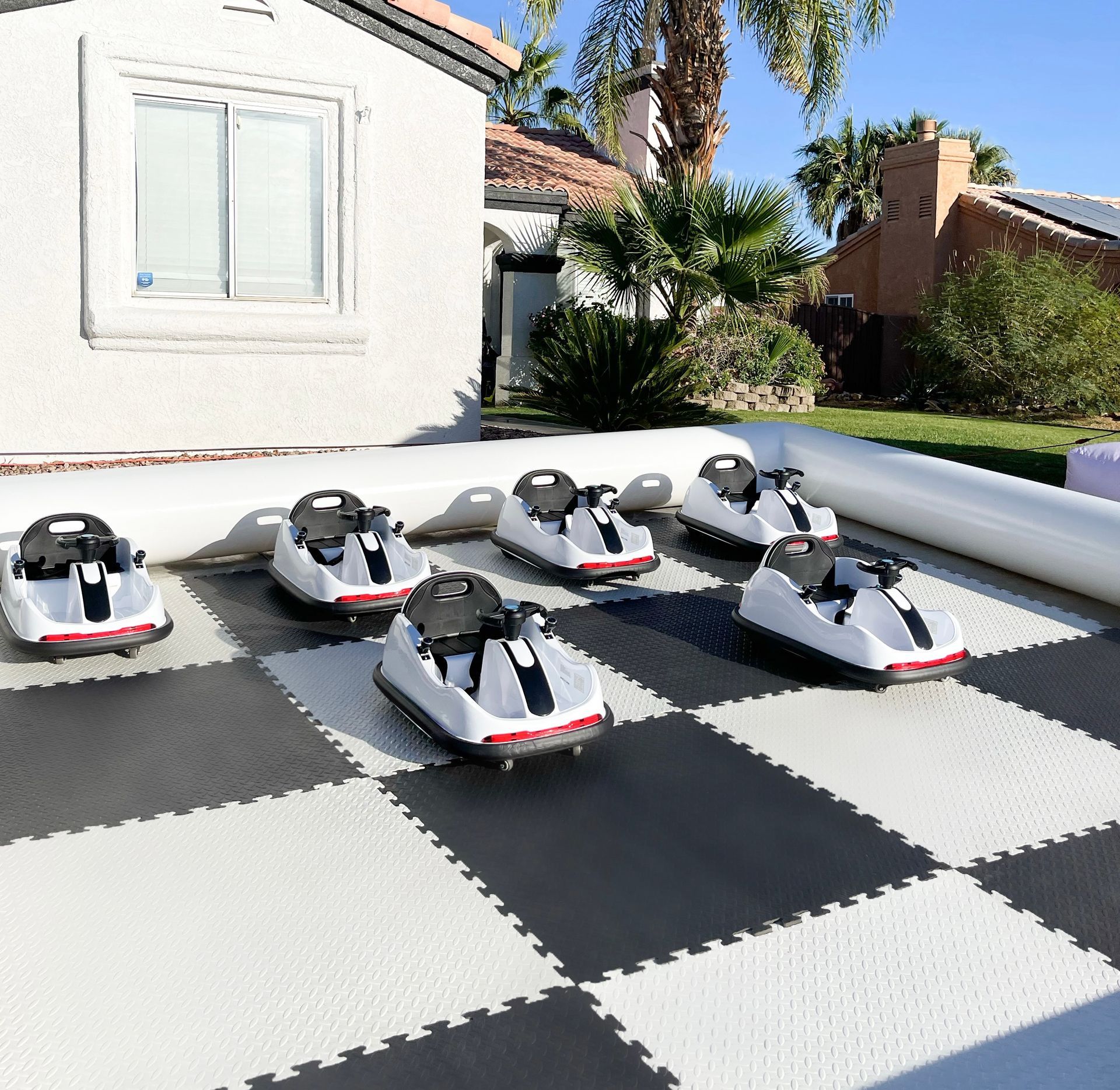Five small gray-and-white ride-on cars lined up on a black-and-white checkerboard patio in a sunny yard