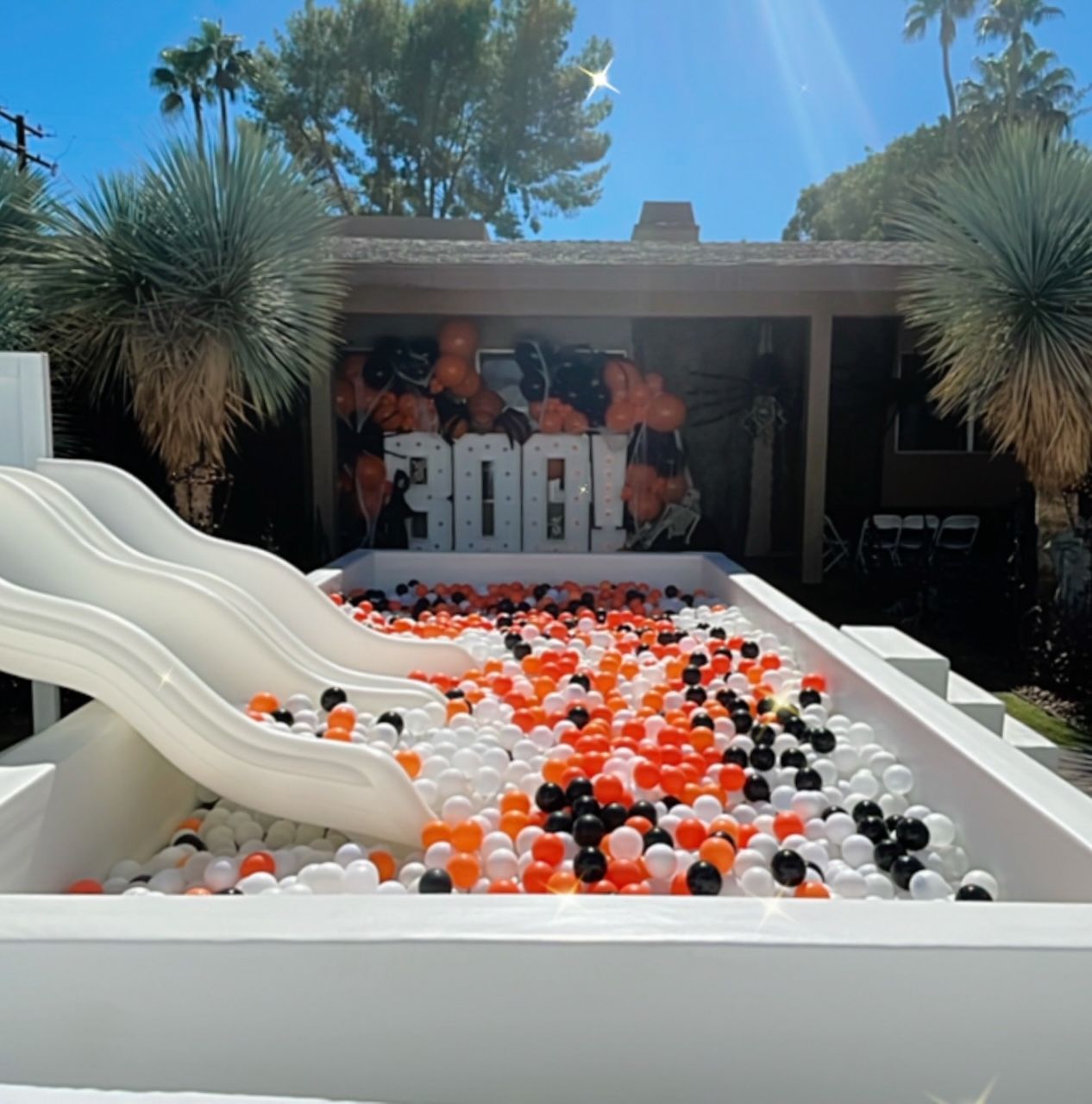 White pool with black, orange, and white balloons beside a slide under a sunny sky