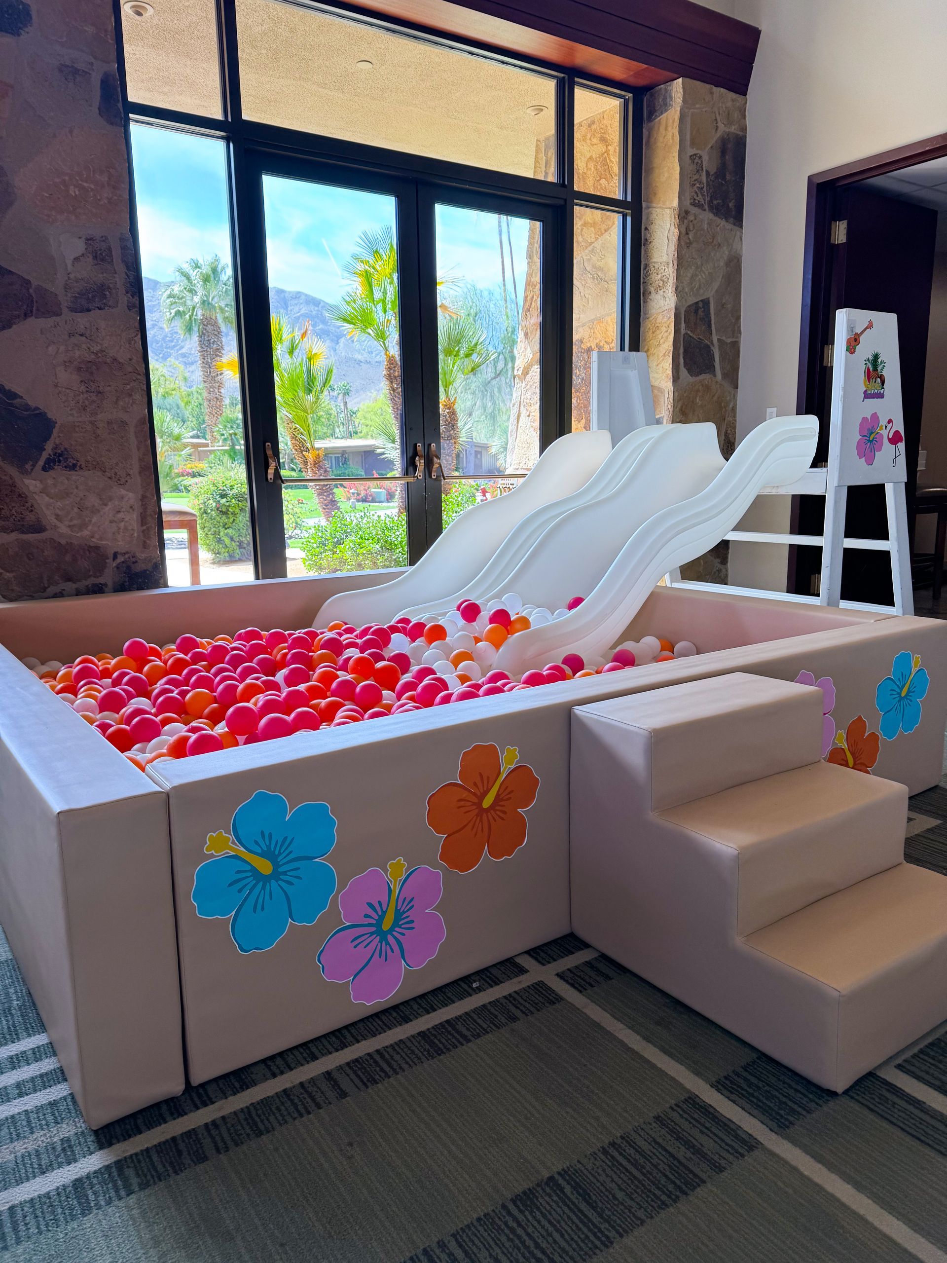 Indoor kiddie ball pit with white slide, pink balls, and flower decorations beside tall windows