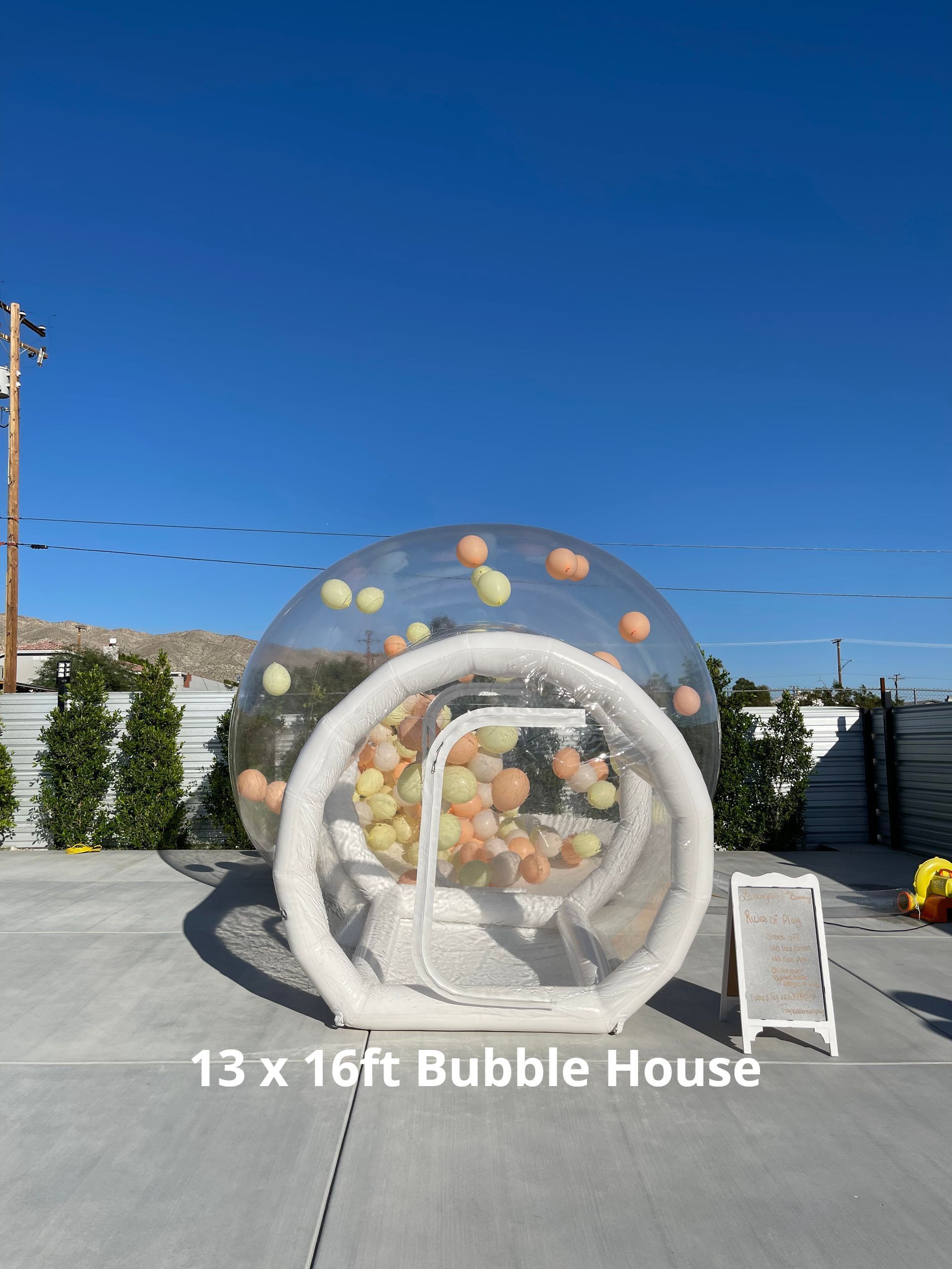 Transparent bubble house display with white entrance and colorful pillows, labeled “13 x 16ft Bubble House”