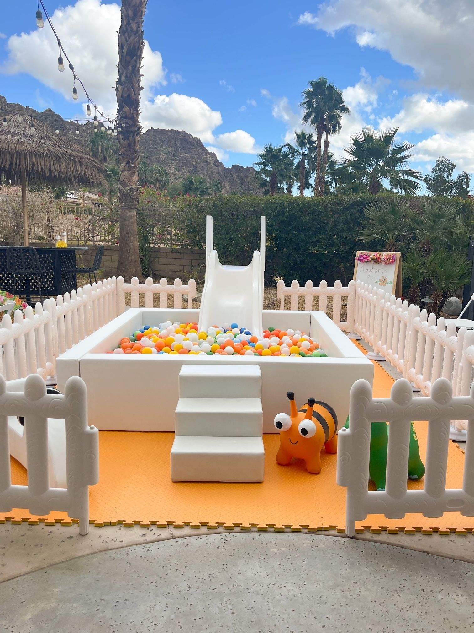 White fenced play area with a small slide, colorful balls, and cartoon animal toys outdoors