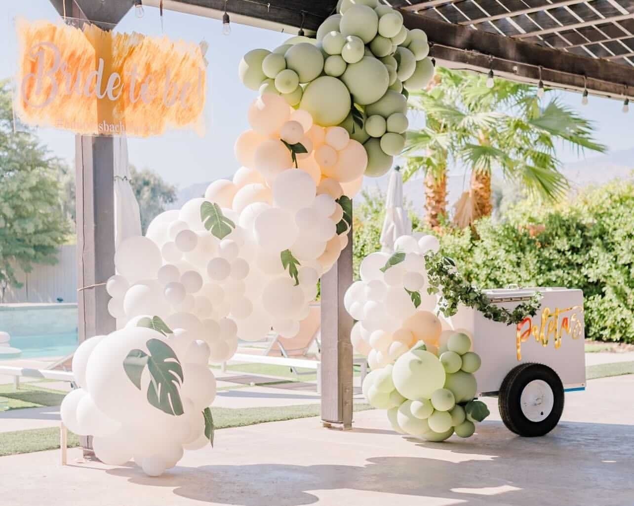 White and green balloon arch at an outdoor party setup with a decorated cart and tropical leaf decor