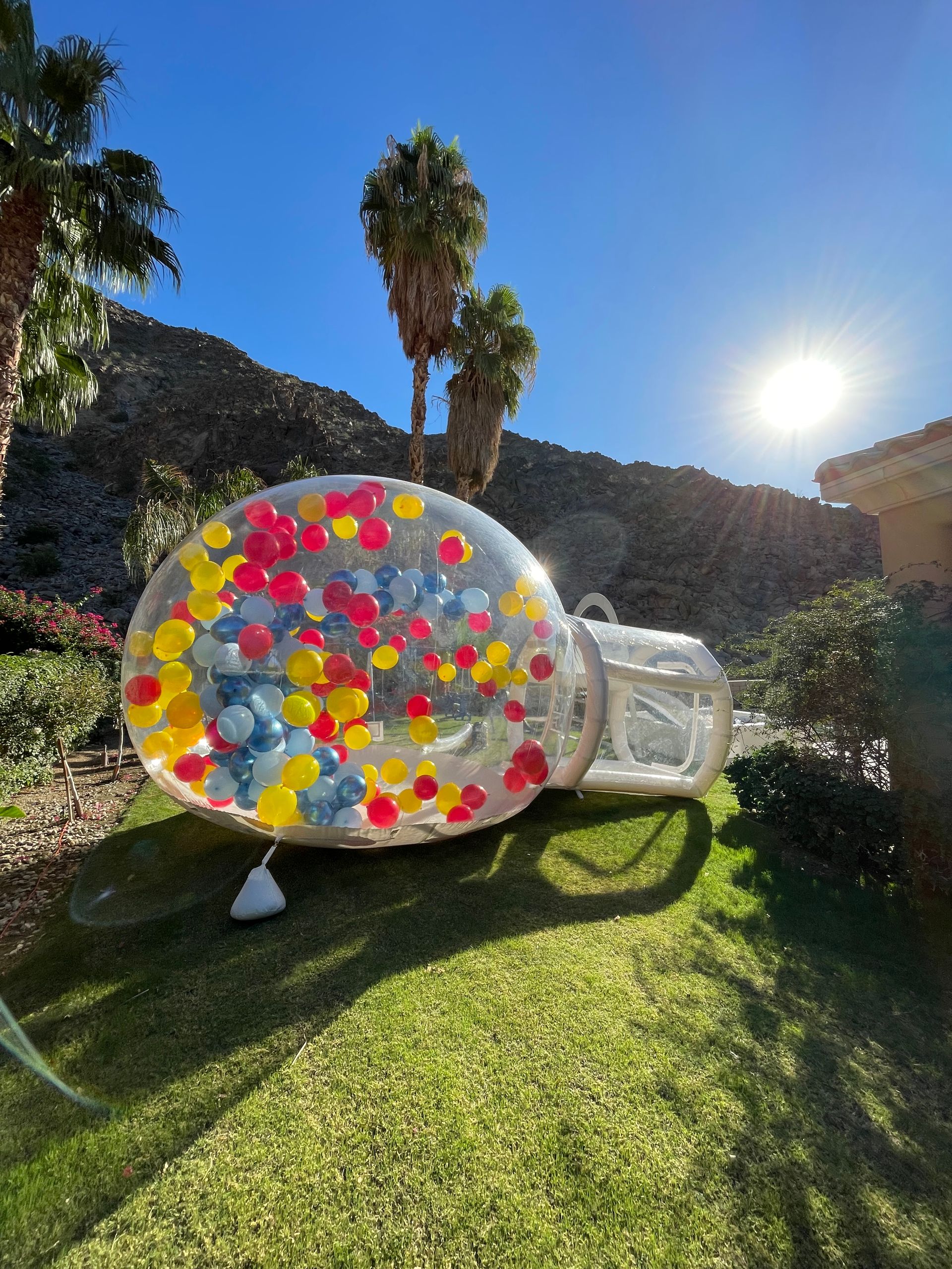 Clear inflatable ball with colorful dots on a sunny lawn, with palm trees and hills in the background.