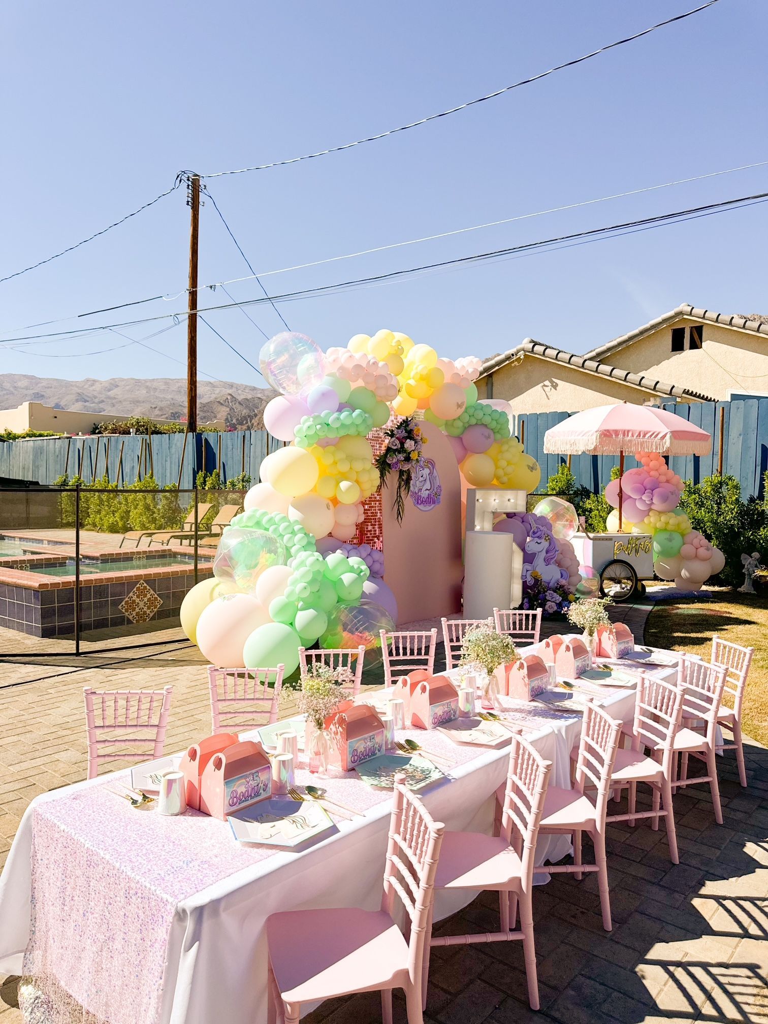 Pastel balloon-decorated outdoor party table with pink chairs and floral accents under a clear sky