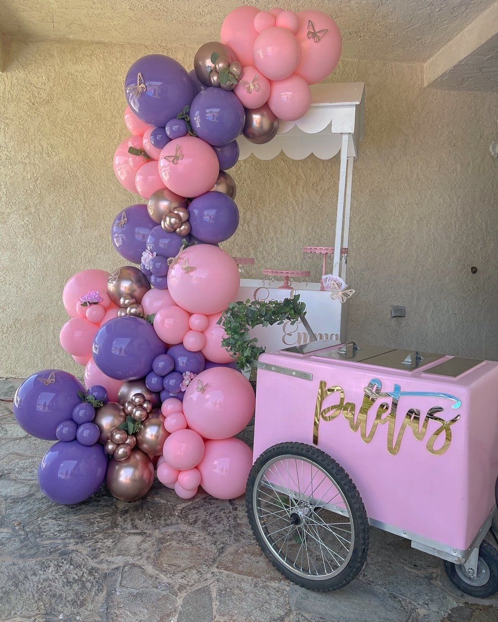 Pink and purple balloon arch beside a pink cart with gold “Baptism” sign in a home entryway