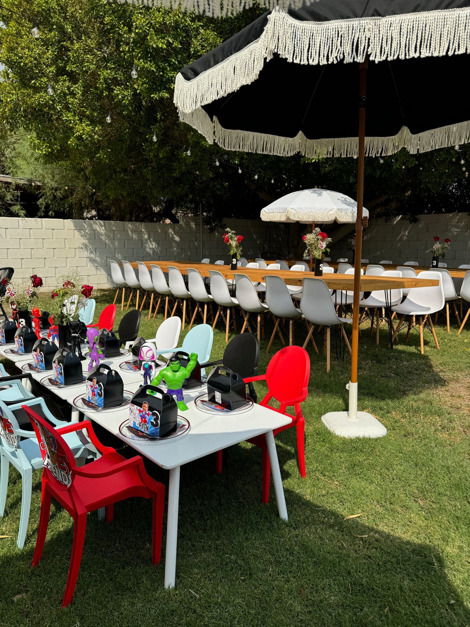 Outdoor event setup with white table, red chairs, and long banquet table under white umbrellas on a lawn.