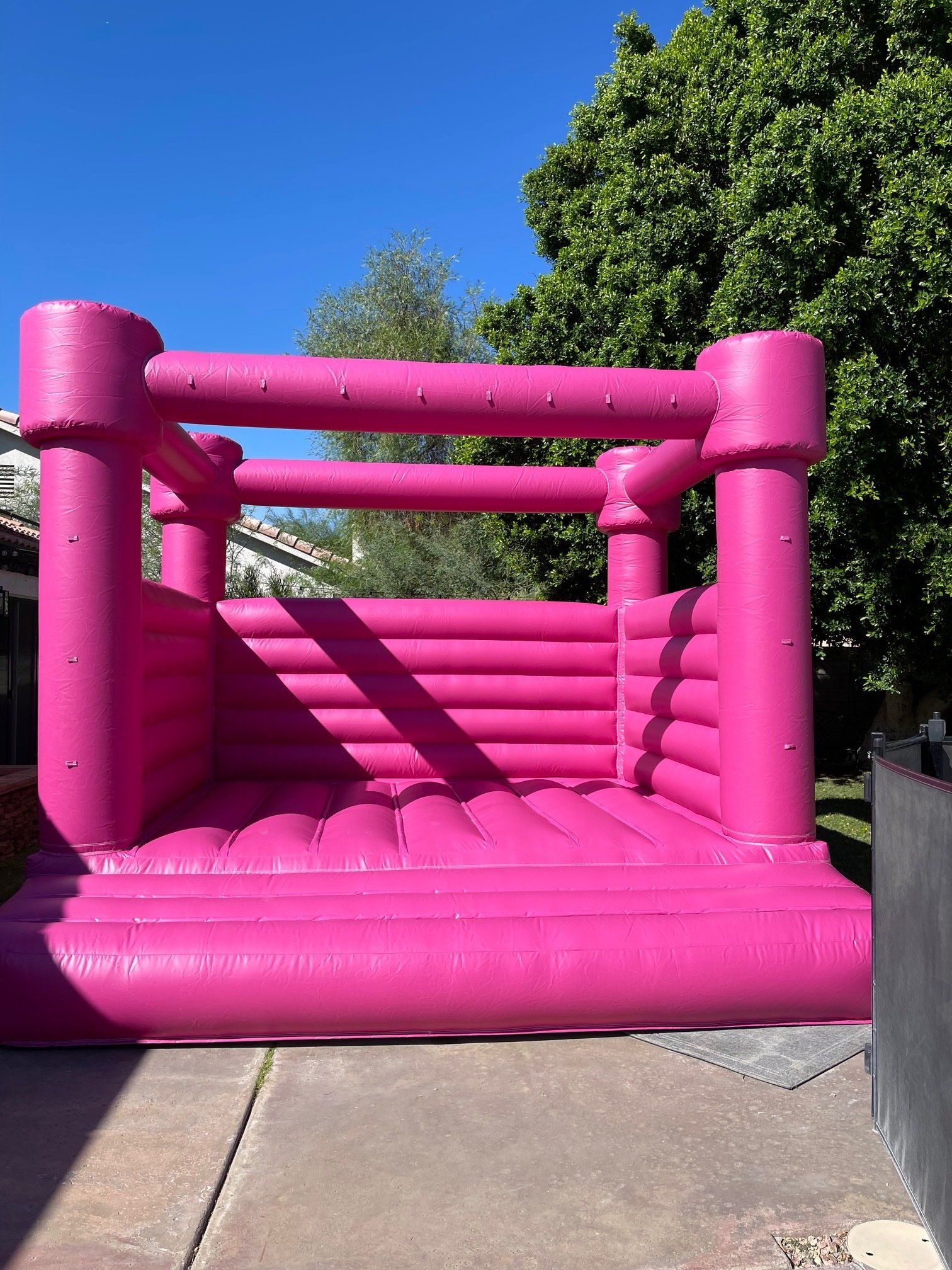 Bright pink inflatable bounce house on a sidewalk, with trees and blue sky behind it.