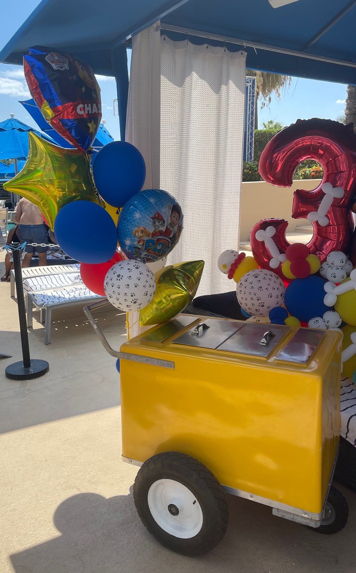 Yellow party cart with blue, red, and gold balloons on a sunny patio