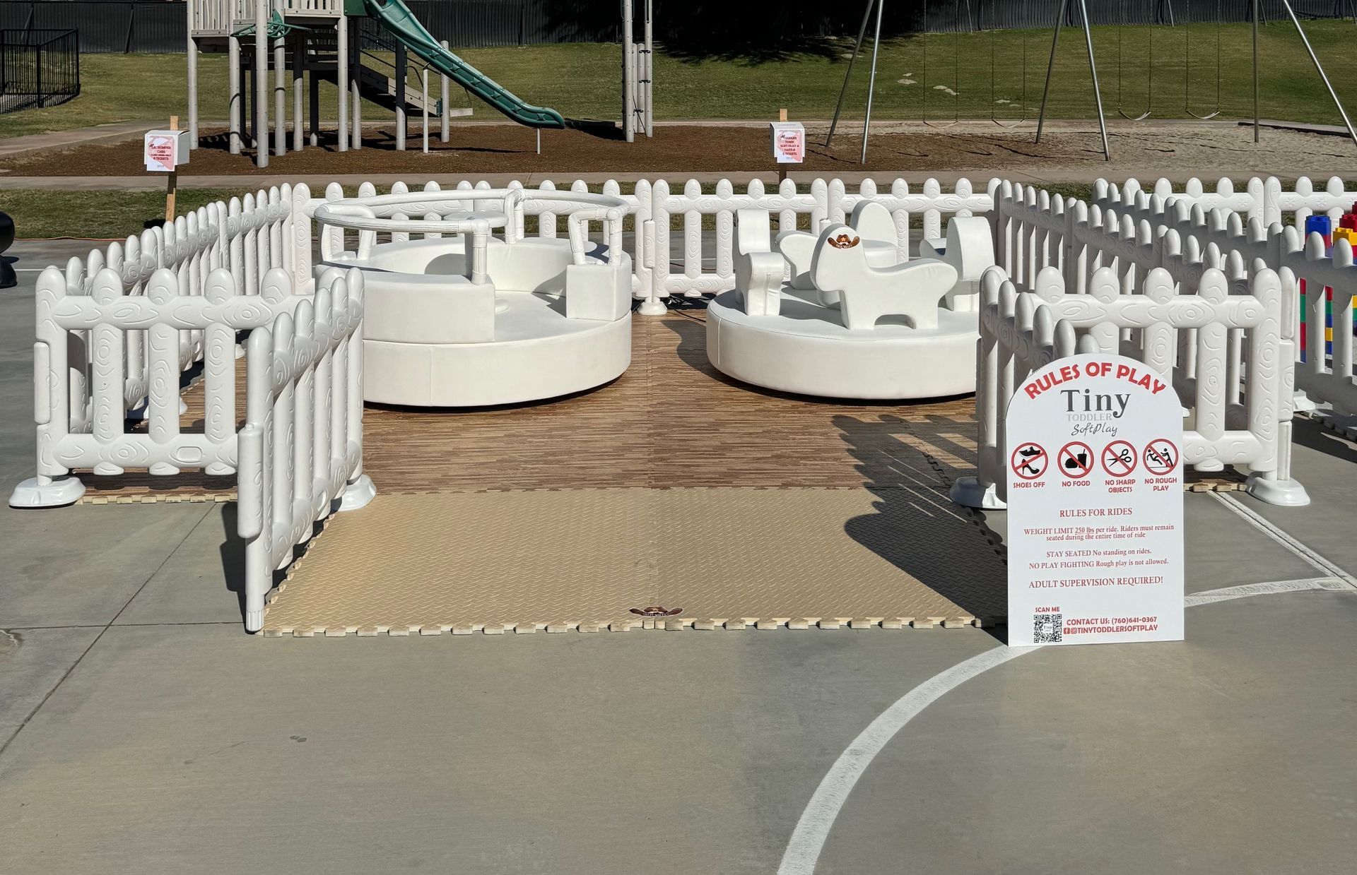 White fenced outdoor play area with round sand tables and a posted sign on a paved surface