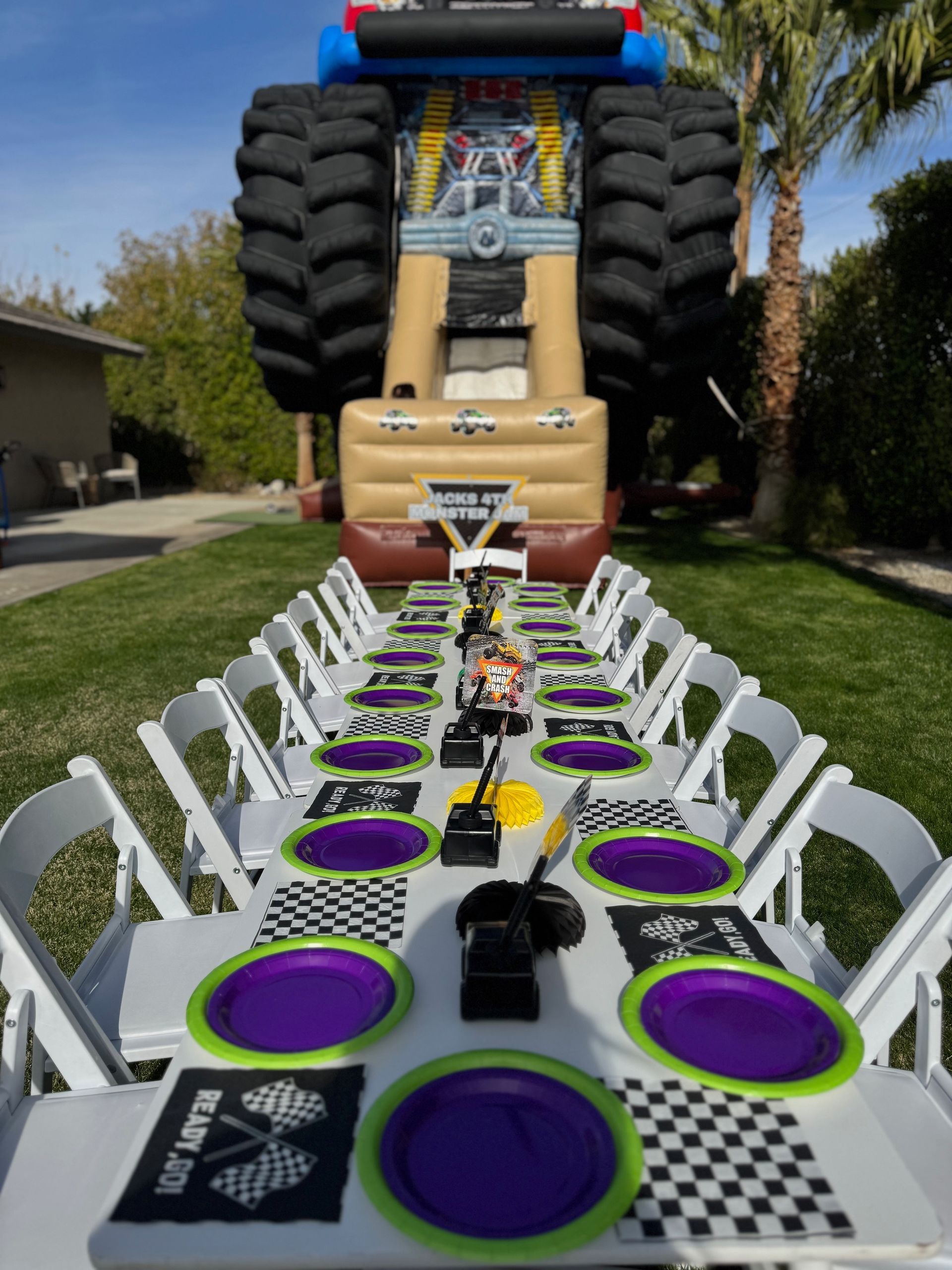 Outdoor table decorated with purple plates and checkered napkins, viewed from a massive truck at the end of the lawn