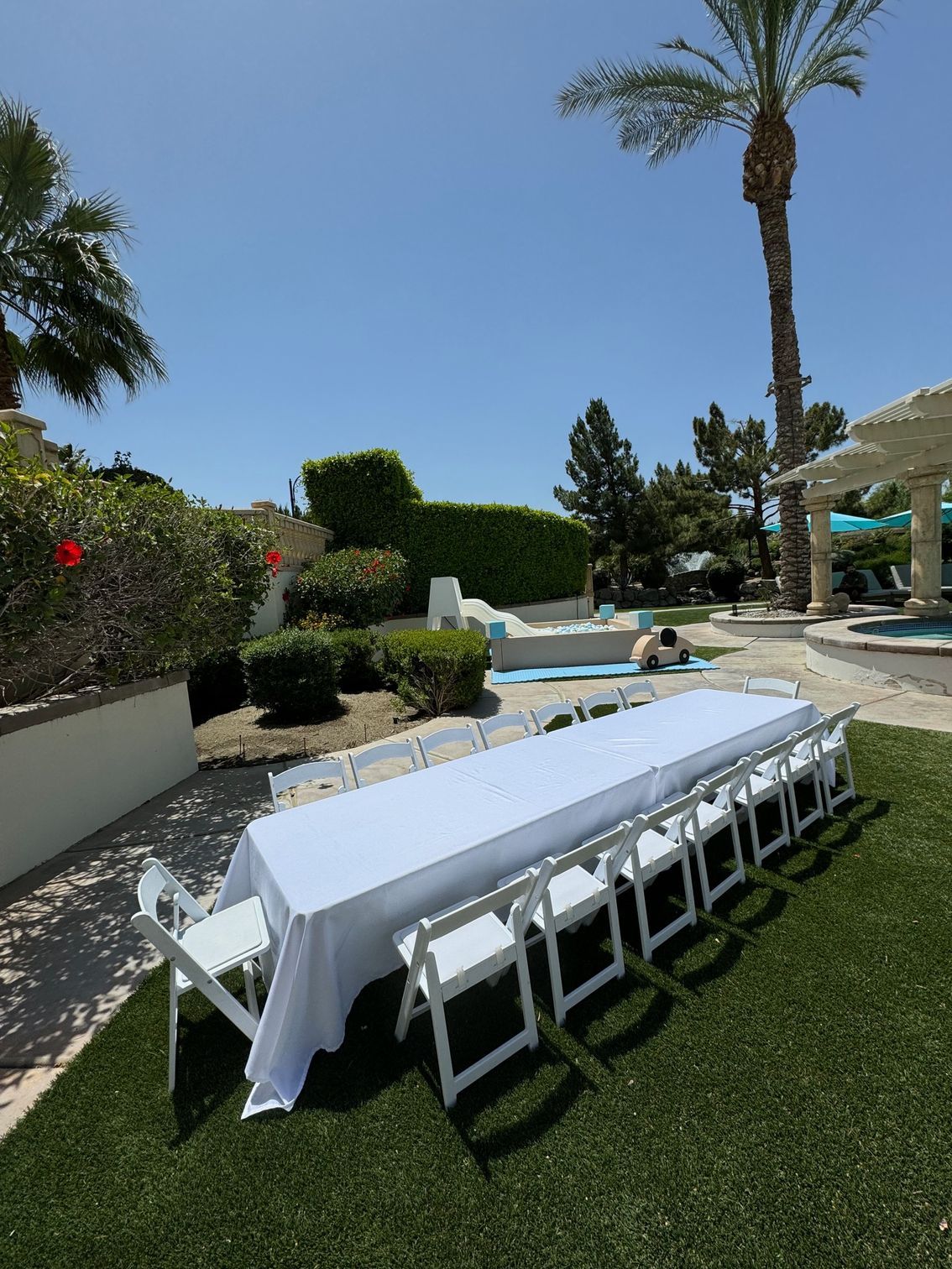Outdoor patio with a long white table and chairs on a lawn beside a pool, under palm trees and clear sky