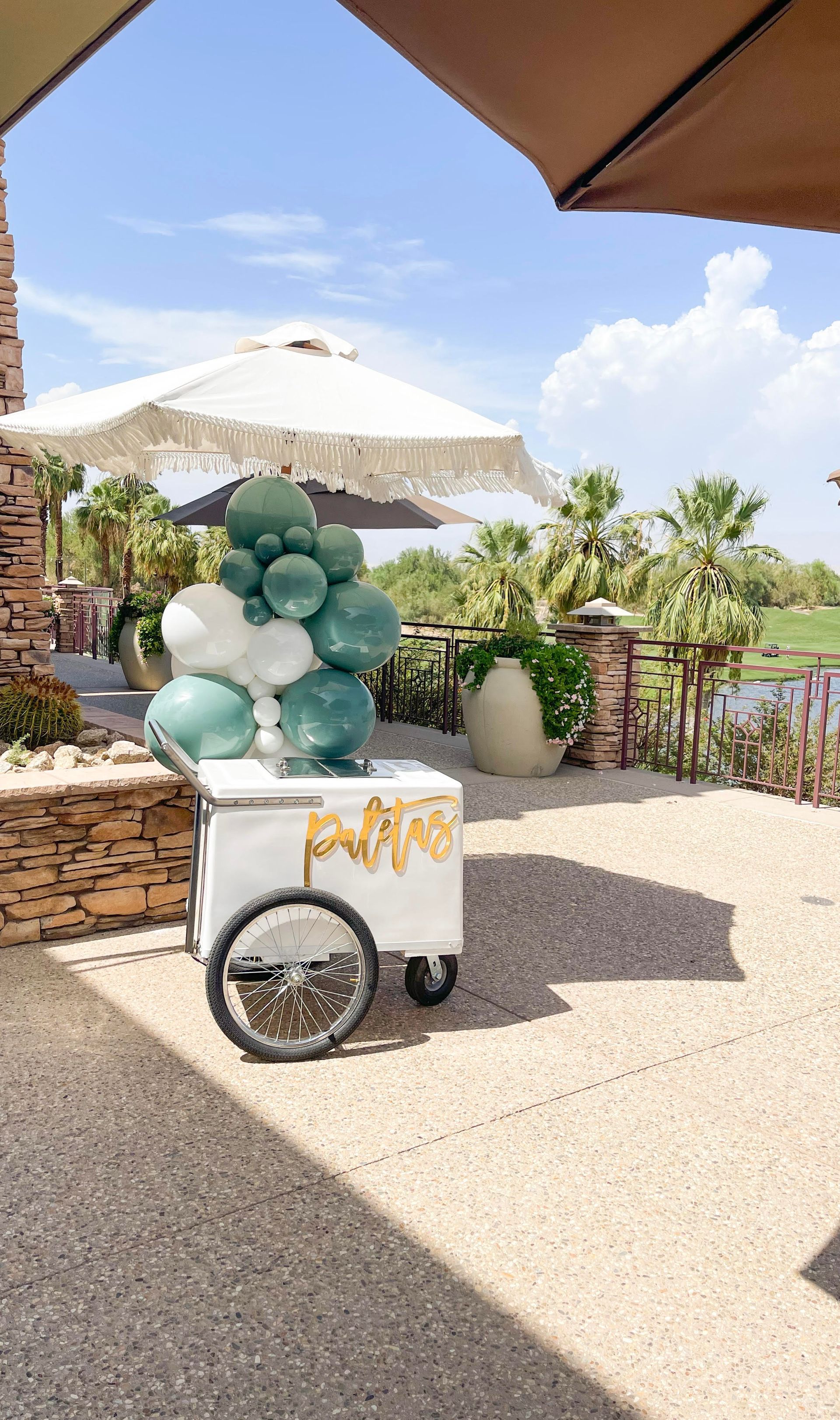 White cart with teal balloon sculpture on a sunny patio near umbrellas and water view