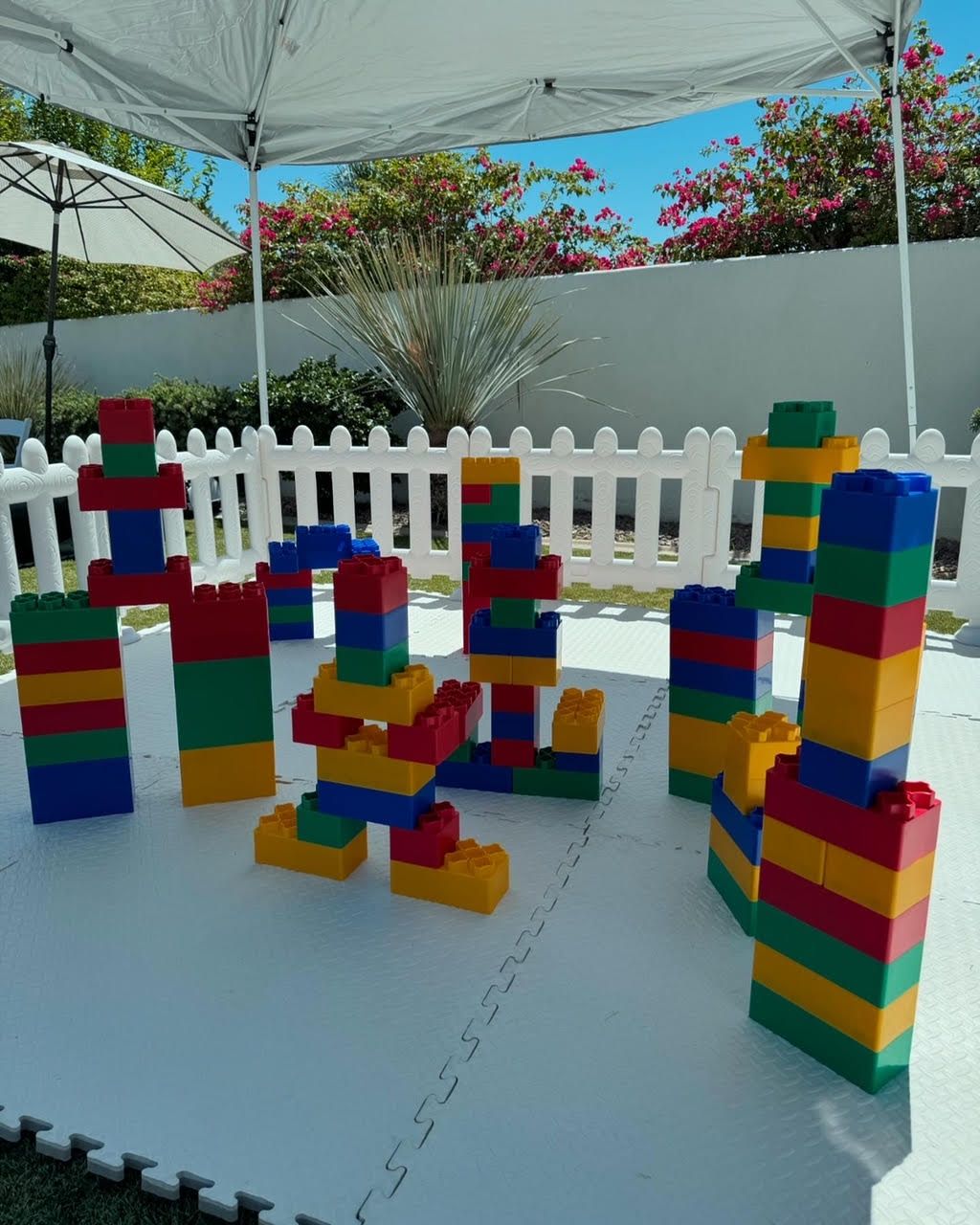 Colorful stacked toy blocks arranged on a white play mat in an outdoor play area with a picket fence.