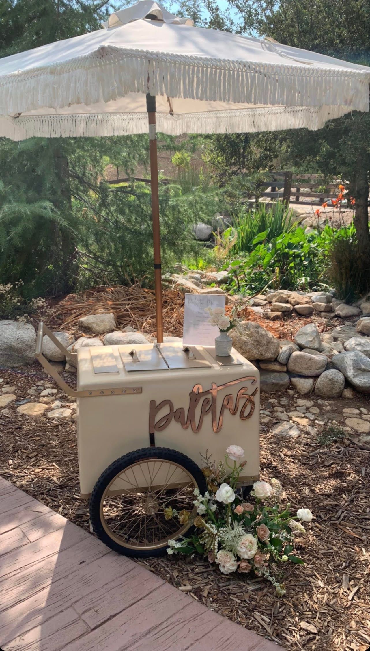 Small outdoor lemonade stand under a white umbrella, with a bicycle wheel sign and greenery behind it.