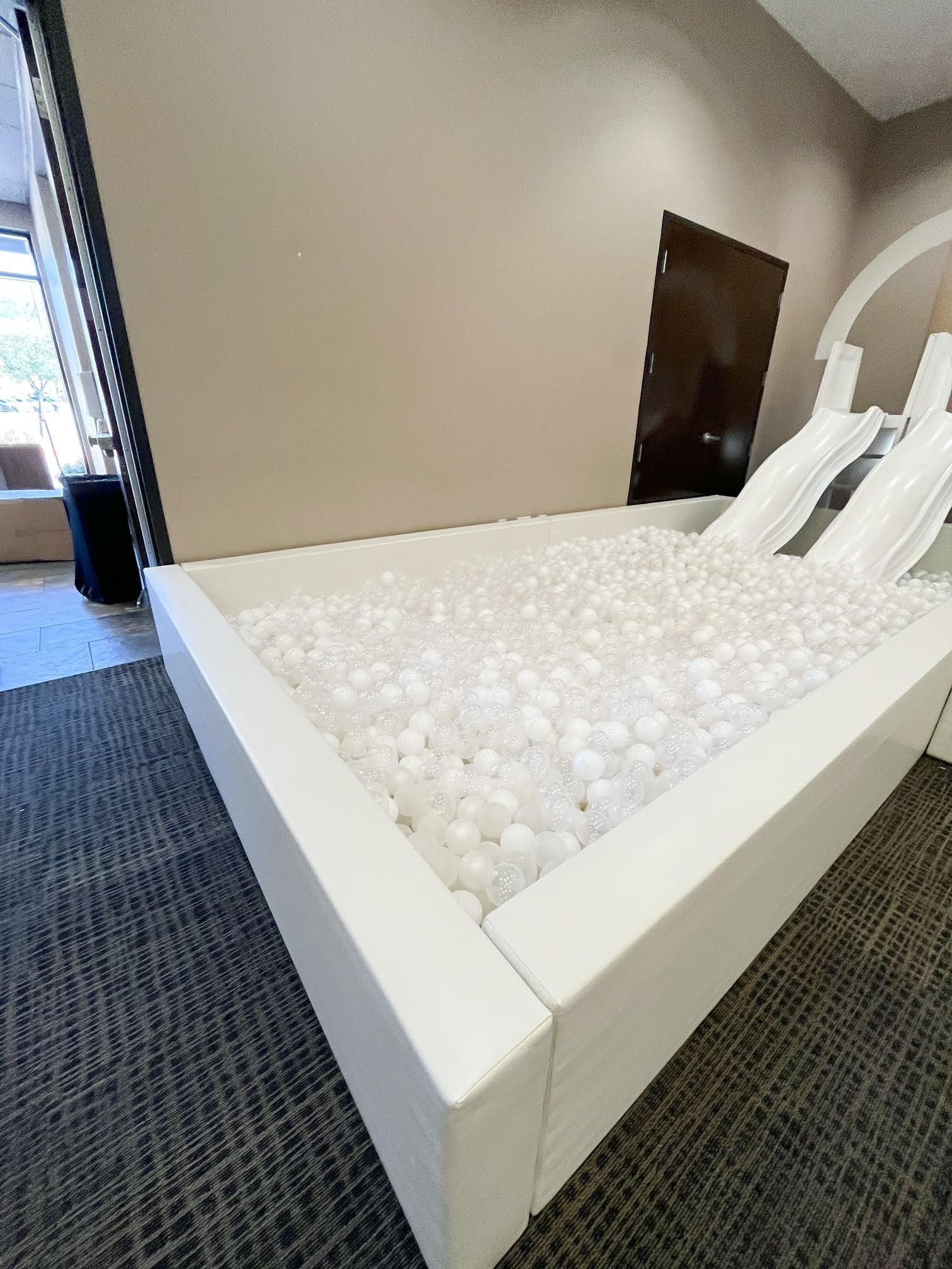 White indoor ball pit with two white slides in a play area next to a carpeted hallway