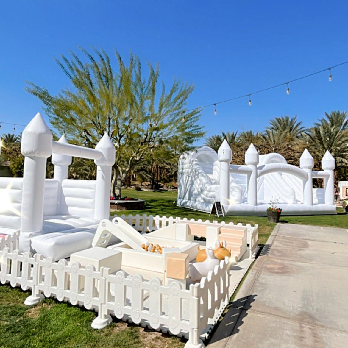 White inflatable bounce houses and playpen on a sunny lawn with palm trees and a driveway