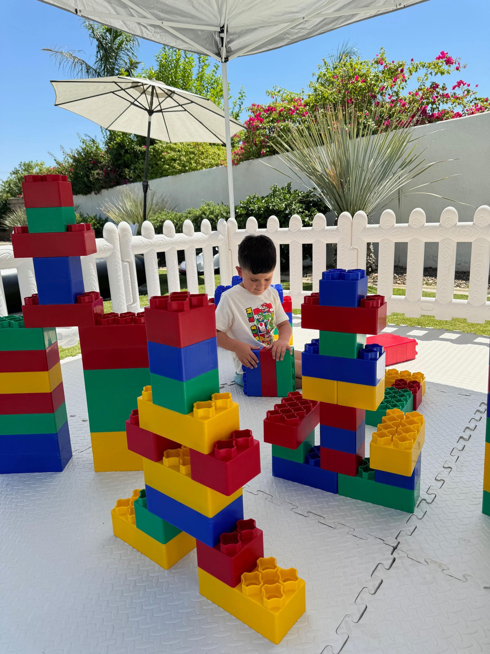 Child playing with colorful blocks in a sunny outdoor play area with umbrellas and a white fence