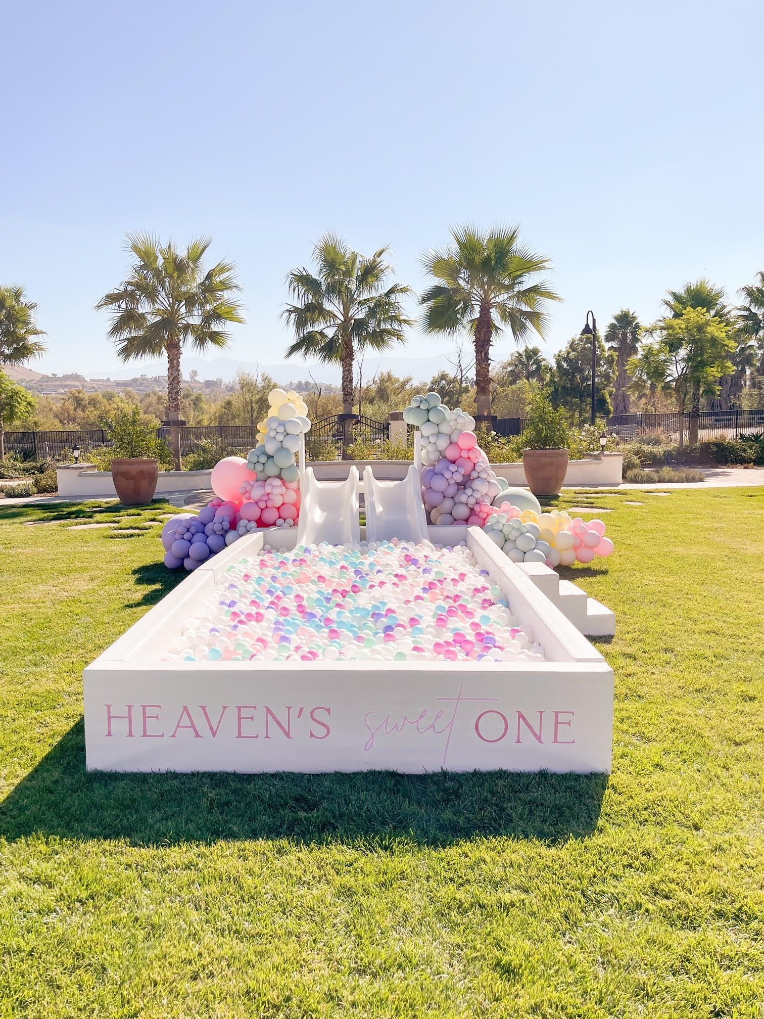 Pastel dessert display on a pink platform labeled “HEAVEN’S ONE” in a sunny garden with palm trees.