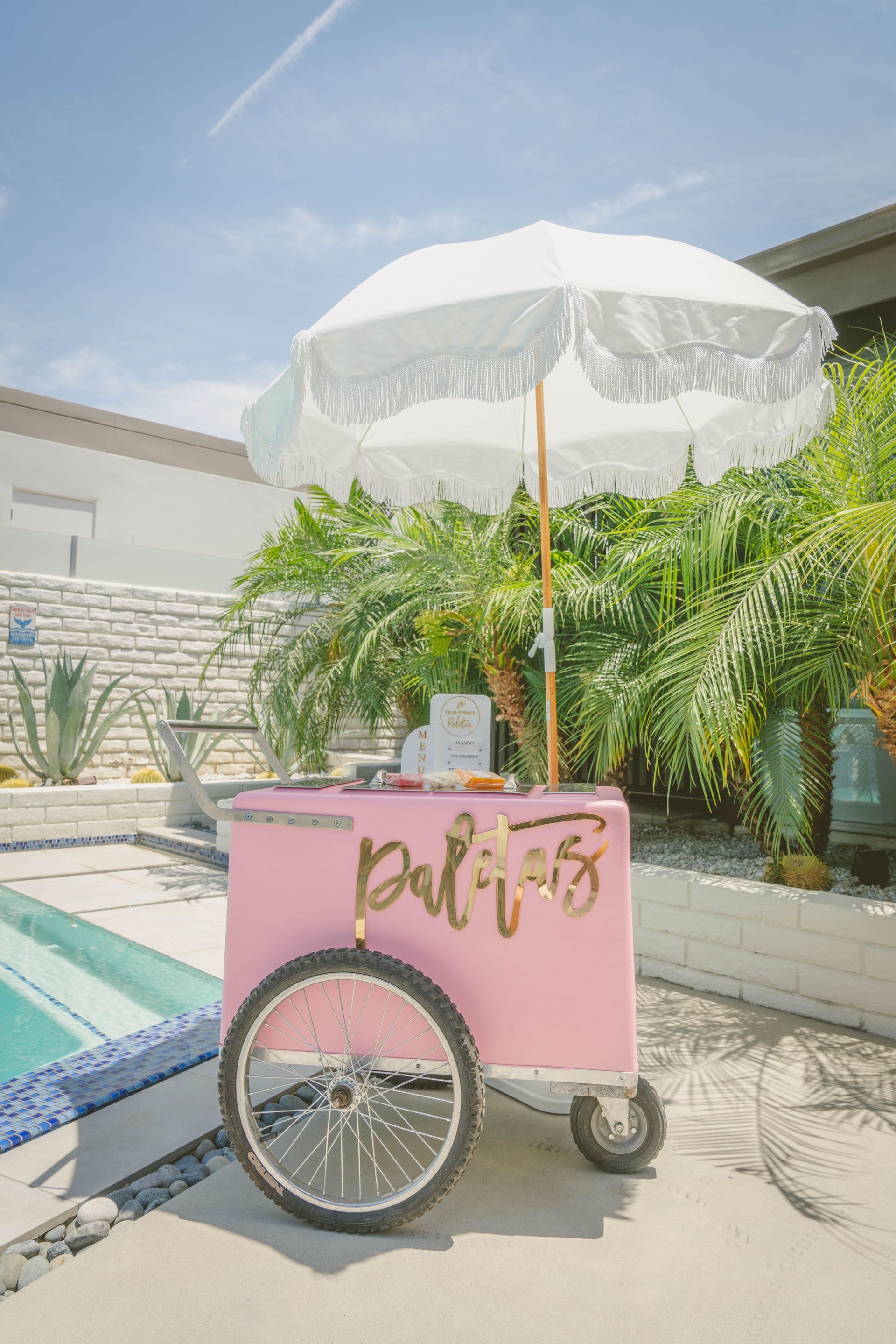 Pink drink cart with a white umbrella and bicycle wheel in a sunny courtyard