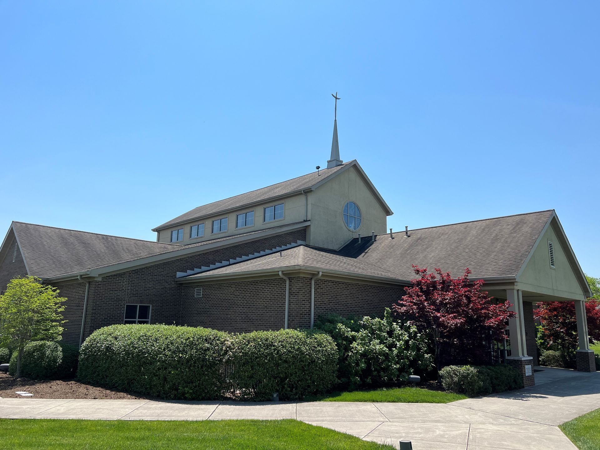 Single-story church with a steeple, tan roof, and manicured shrubs under a clear blue sky