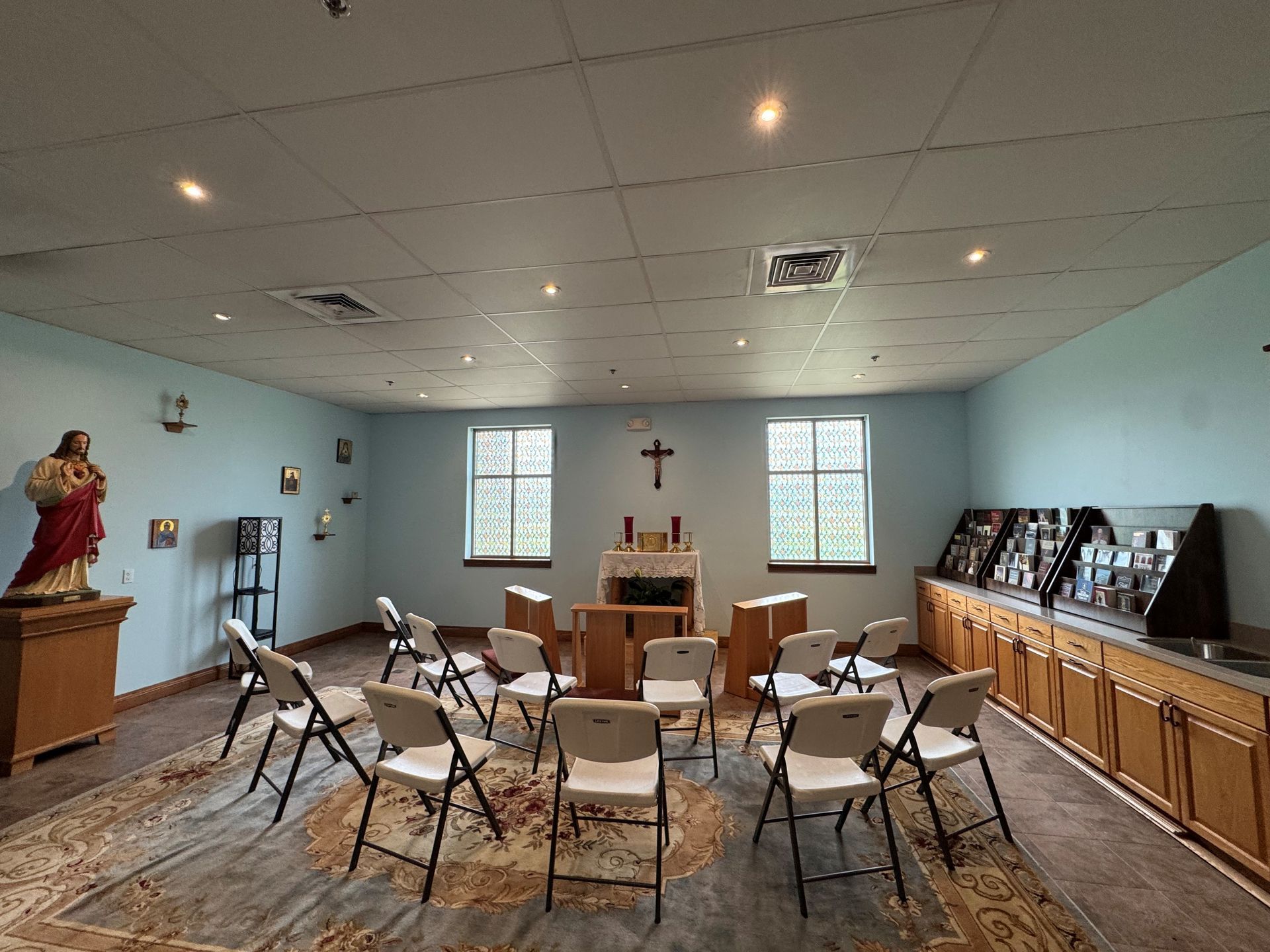 Small chapel with rows of folding chairs facing an altar and crucifix, pale blue walls and cabinets.