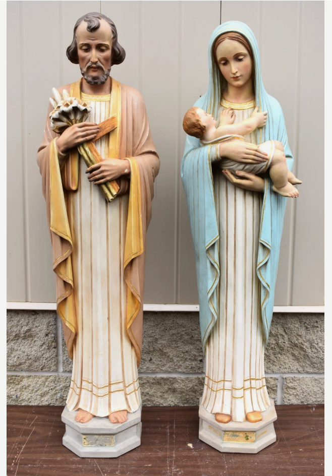 Two religious statues in robes holding flowers, standing side by side against a wall.