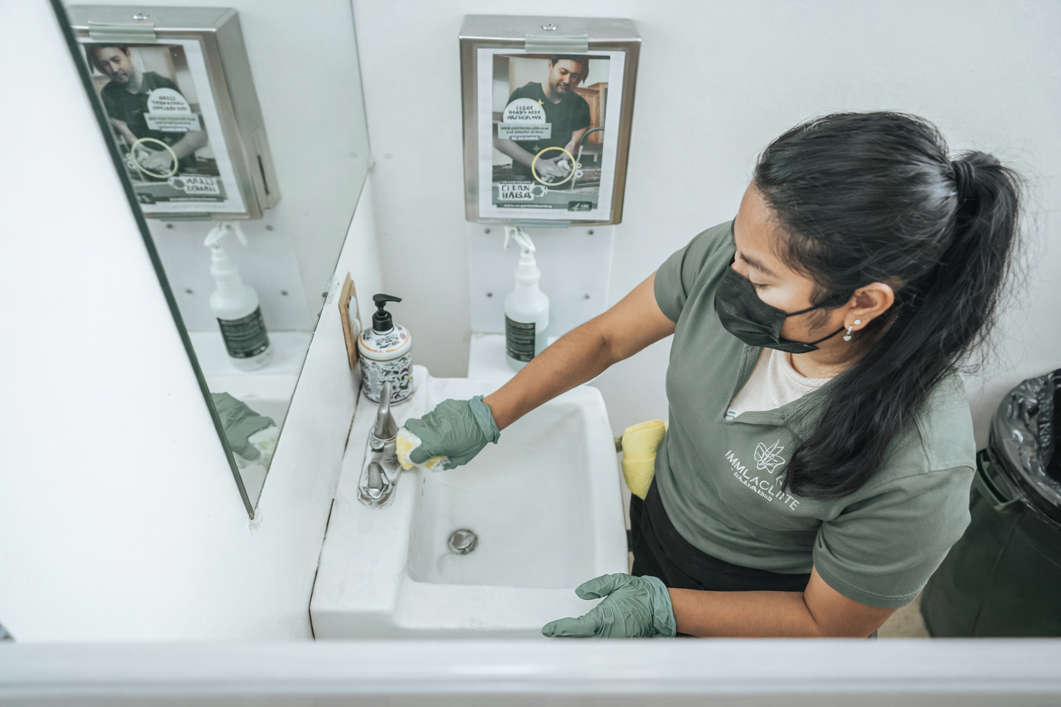 Person cleaning a bathroom sink with a cloth, wearing gloves and a mask, seen in the mirror.