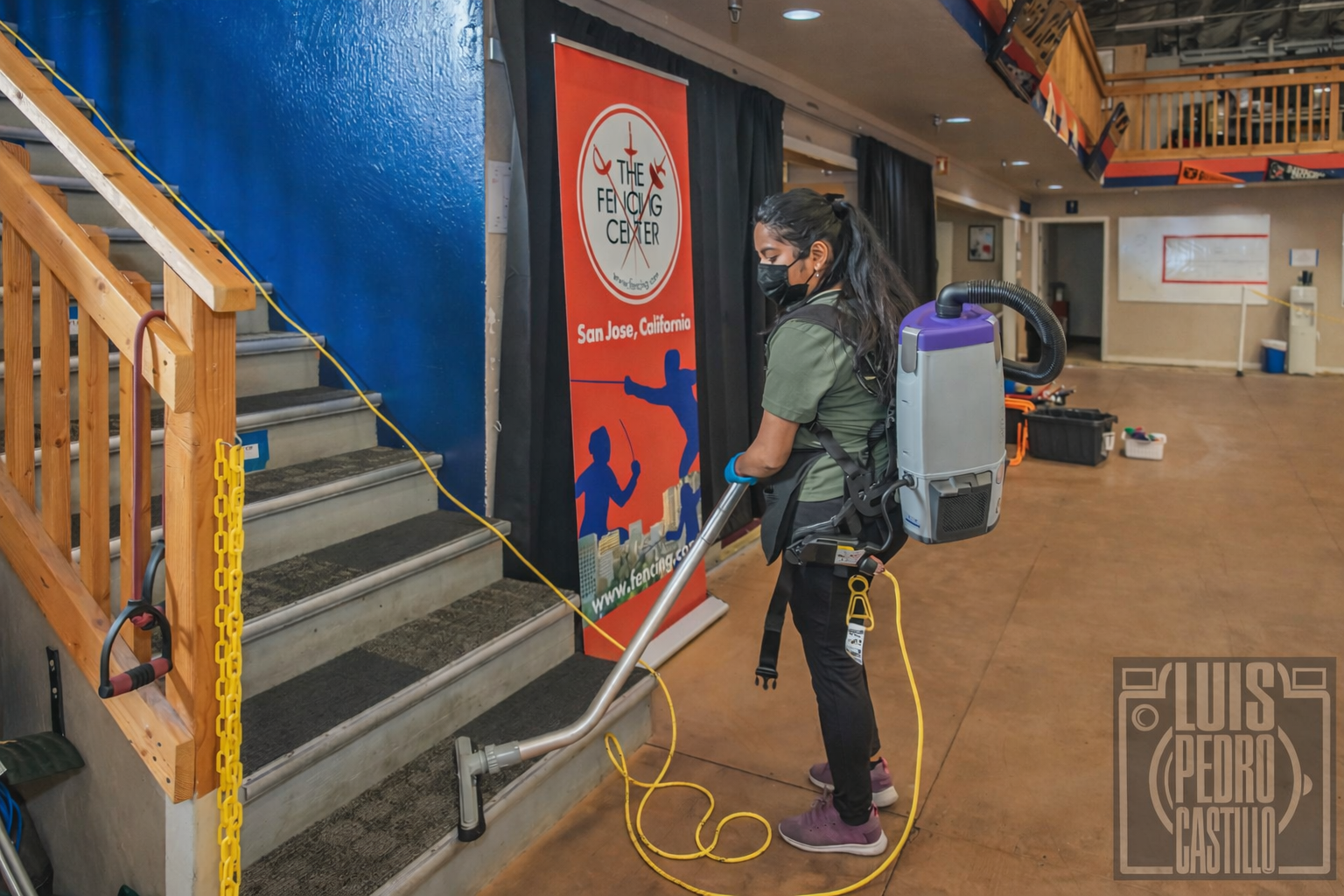 Worker pressure-washes a stairwell inside a building, with a blue wall, orange railings, and cleaning equipment nearby