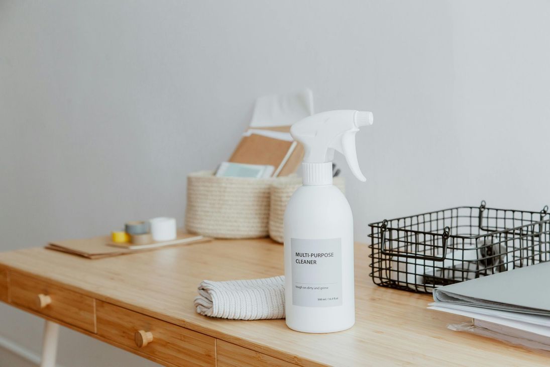 A white spray bottle and a folded cloth sit on a light wood desk.