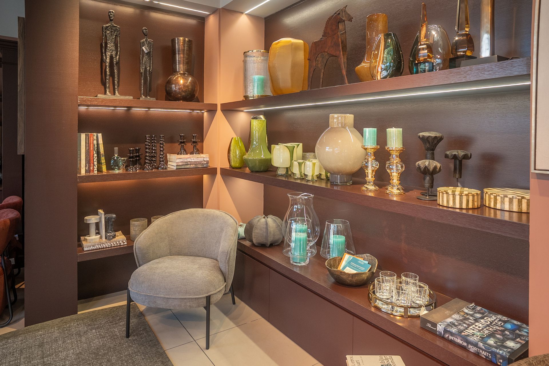 In un ambiente interno accogliente, moderne vetrine da boutique con vasi decorativi, barattoli di vetro e una poltrona grigia.